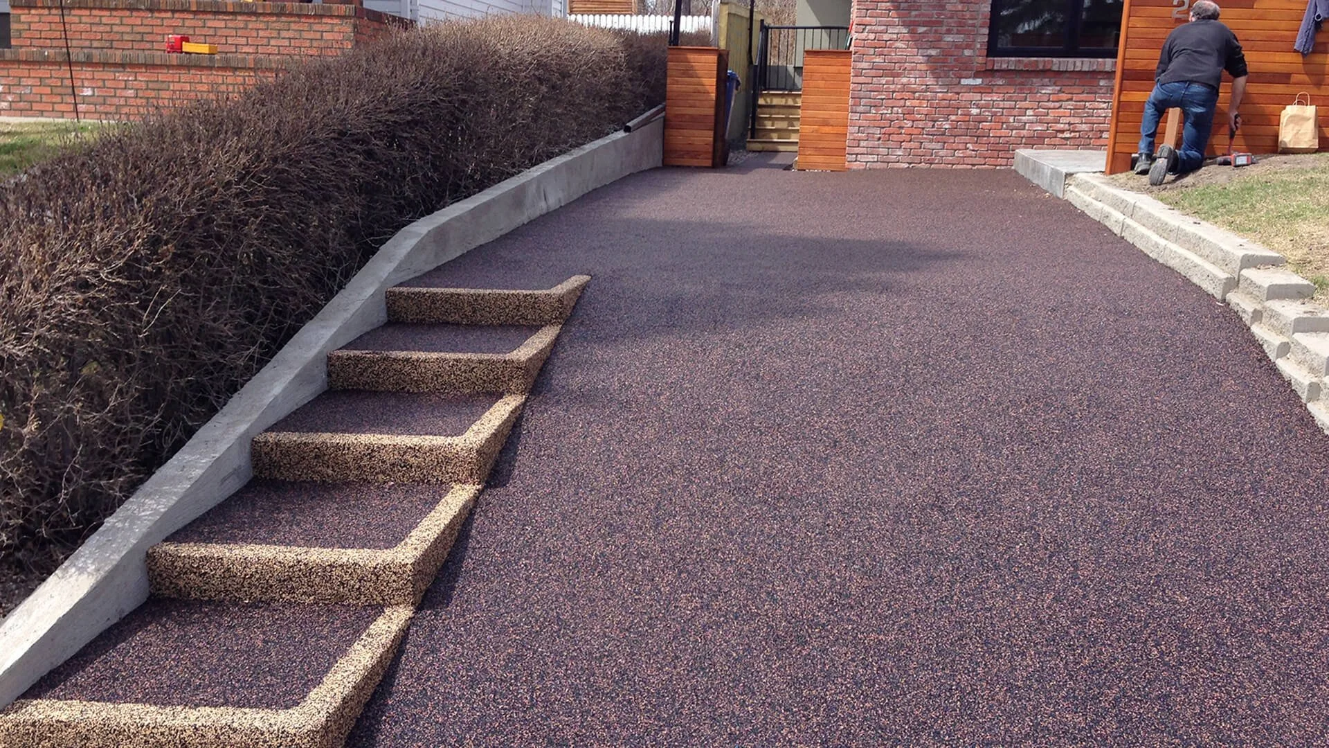 A driveway with newly poured asphalt, a set of three concrete steps, a hedge on the left side, a brick house with a wooden section on the right, and a man kneeling and working on the wooden section of the house.