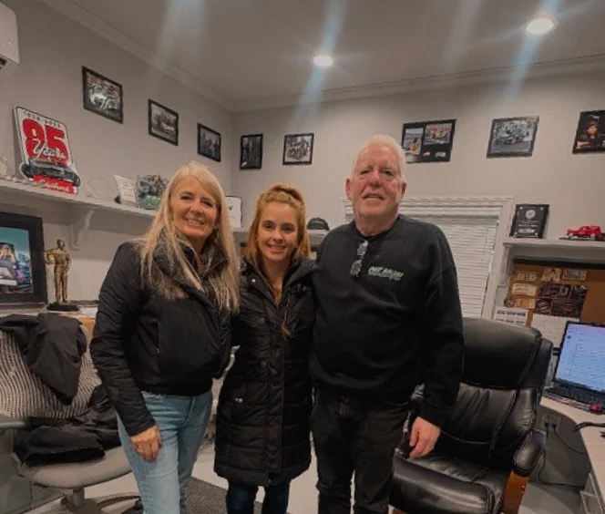 Three people, two women and one man, standing and smiling in an office with framed photos and memorabilia on the walls
