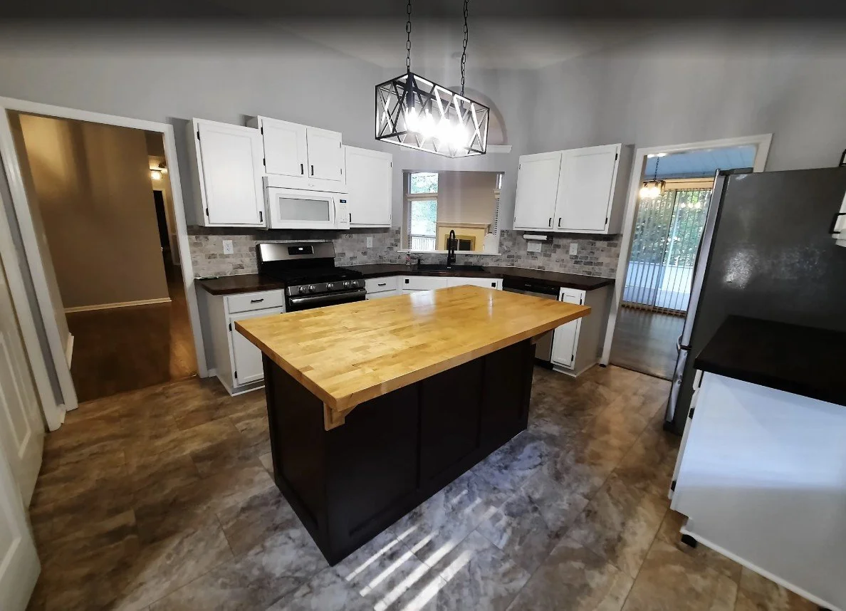 Kitchen with white cabinets, black countertops, a wooden kitchen island, and a black stove. Hanging light fixture above island, sliding door leading outside, window behind sink, and gray tile backsplash.
