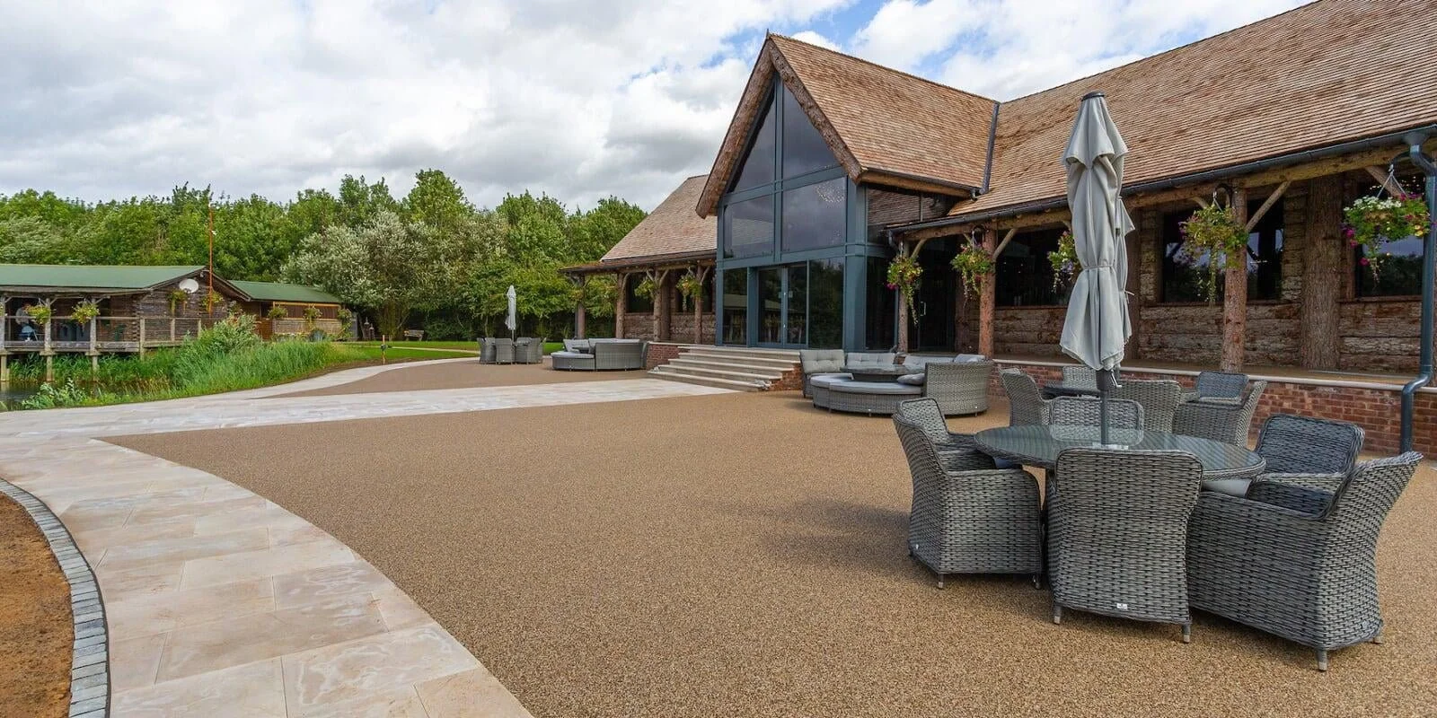 Outdoor patio with wicker furniture, a glass-top table with an umbrella, and a large building with glass windows and wooden exterior, surrounded by lush green trees.