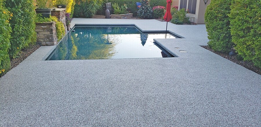 Backyard with a concrete pool deck surrounding a rectangular swimming pool, with lush green bushes and trees on both sides, a patio umbrella, and a house in the background.