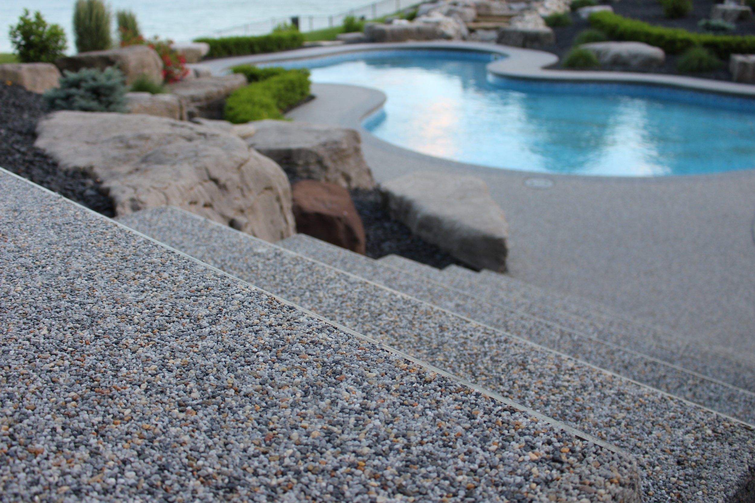 Close-up of a gravel pathway leading to a swimming pool, with rocks, plants, and water in the background.