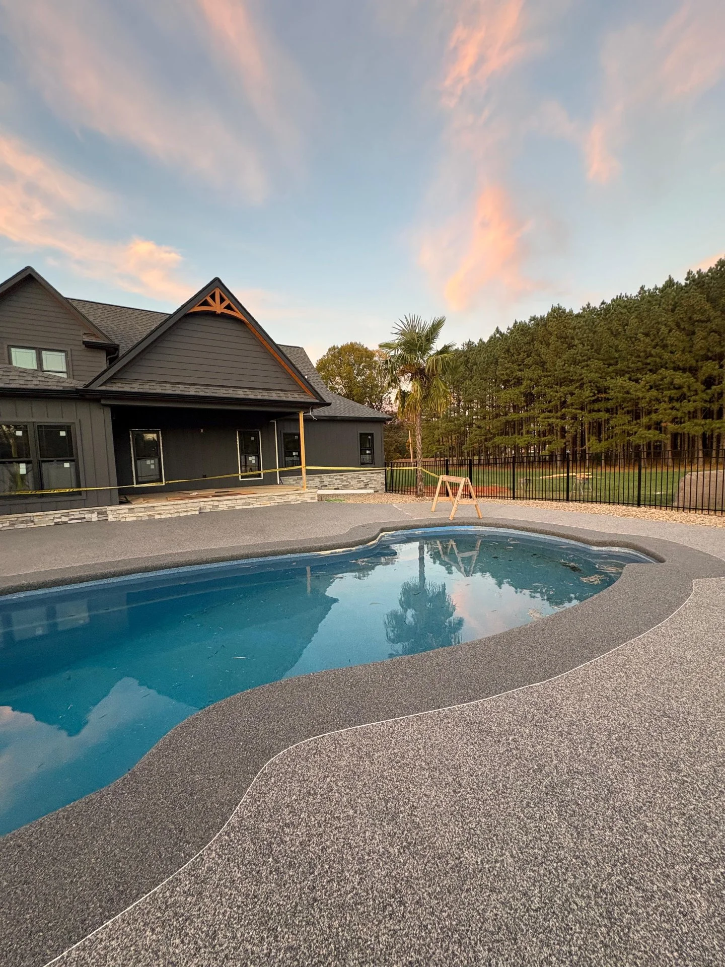 A backyard with a swimming pool, a house with dark gray siding, a wooden fence, and trees in the background under a partly cloudy sky at sunset.