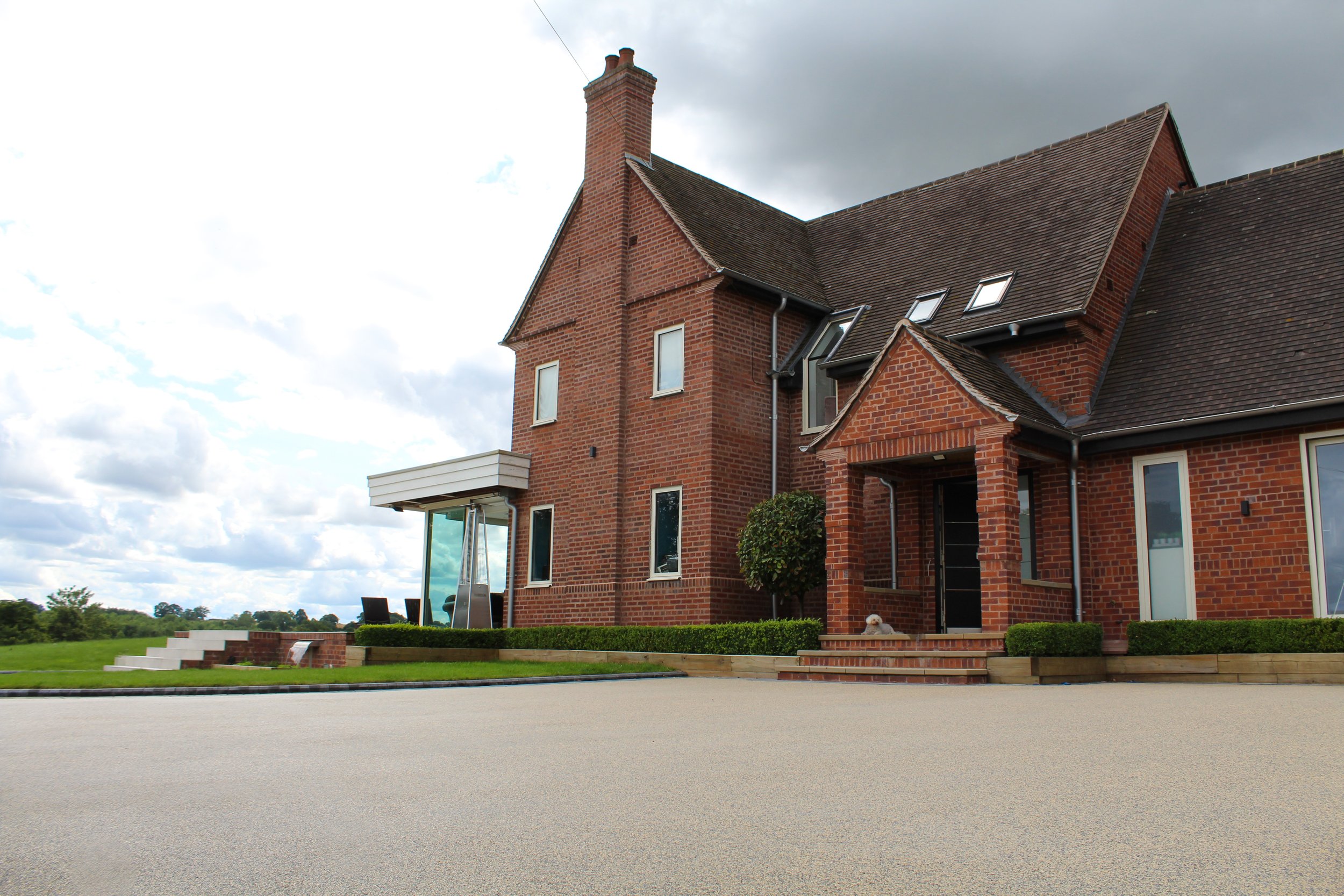 A large red brick house with a steeply pitched roof and multiple windows, situated in a grassy area with a sidewalk in the foreground and a partly cloudy sky overhead.