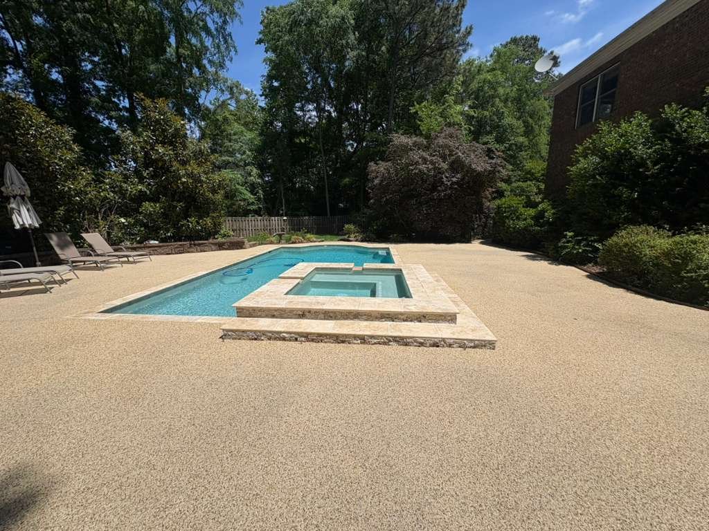 A backyard swimming pool with a small adjacent hot tub, surrounded by a tan-colored concrete deck, with trees and bushes in the background, and a house with windows on the right.