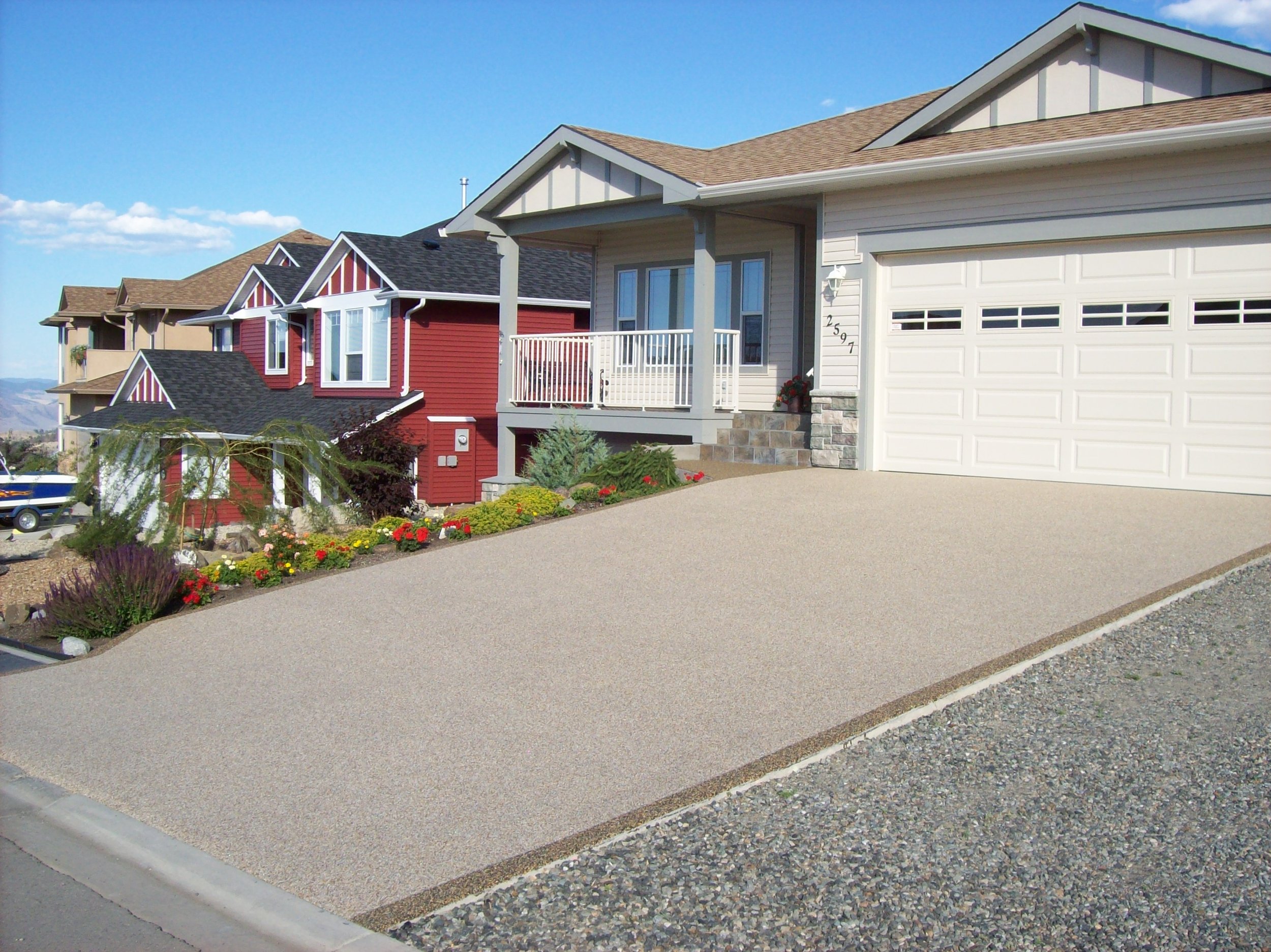 A row of modern houses with different colored facades, a manicured front yard with flowers, and a paved driveway leading to a garage, under a clear blue sky.