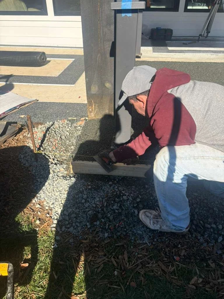 A man in a gray hoodie, maroon jacket, and gray cap is crouching down and working with a trowel on a concrete project outdoors. There is a large metal pole and a building with large windows in the background, with construction materials and tools aro