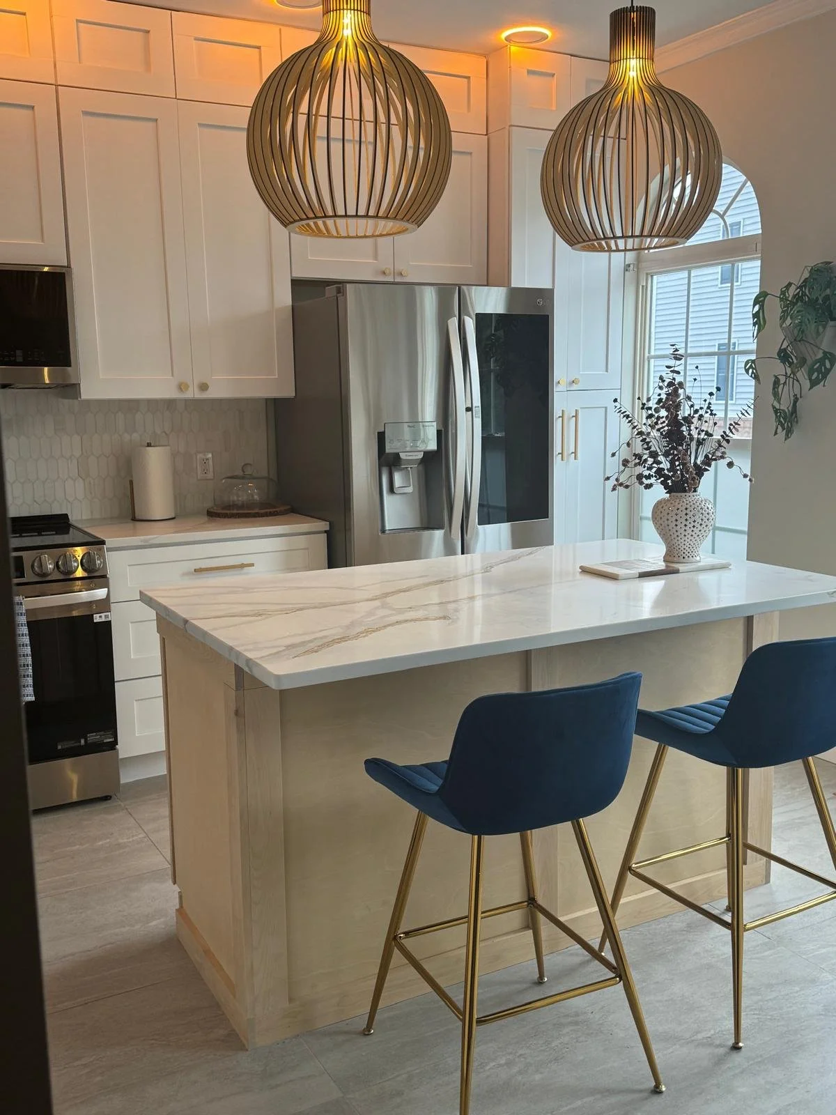 Modern kitchen with white cabinets, stainless steel refrigerator, marble island counter, blue barstools with gold legs, and decorative pendant lights