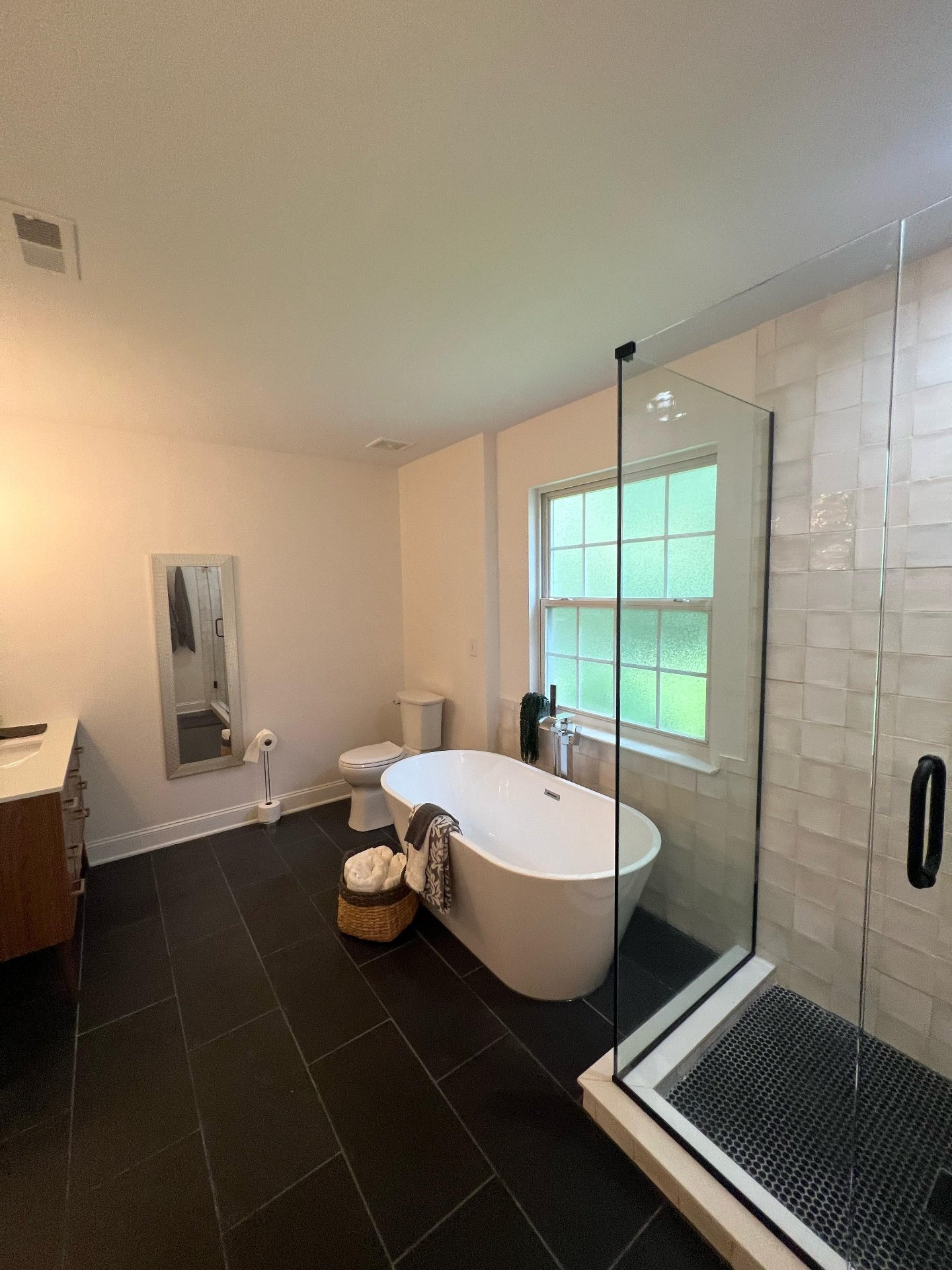 Modern bathroom with a freestanding bathtub, a glass shower enclosure, a toilet, a window with frosted glass, and a small sink vanity. The floor is covered with black tiles, and the walls have a mix of white paint and white subway tiles. There is a b