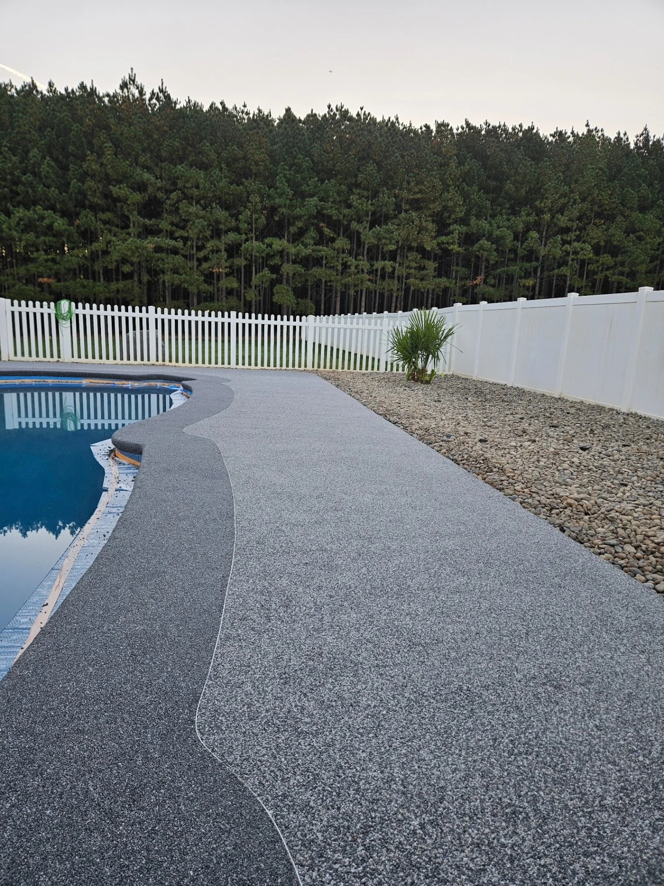 Backyard scene with a kidney-shaped swimming pool, a textured concrete deck, a white vinyl fence, and a wooded area with tall trees in the background.