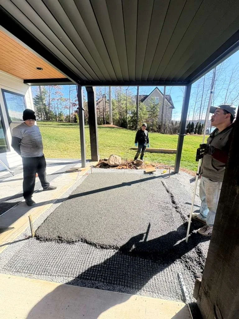People laying down fresh concrete for a patio or sidewalk outside a house, with a grassy yard and trees in the background.
