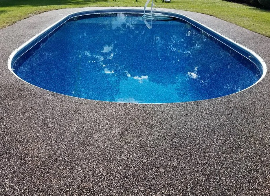 A small blue swimming pool surrounded by a textured gray surface with grass and trees in the background.