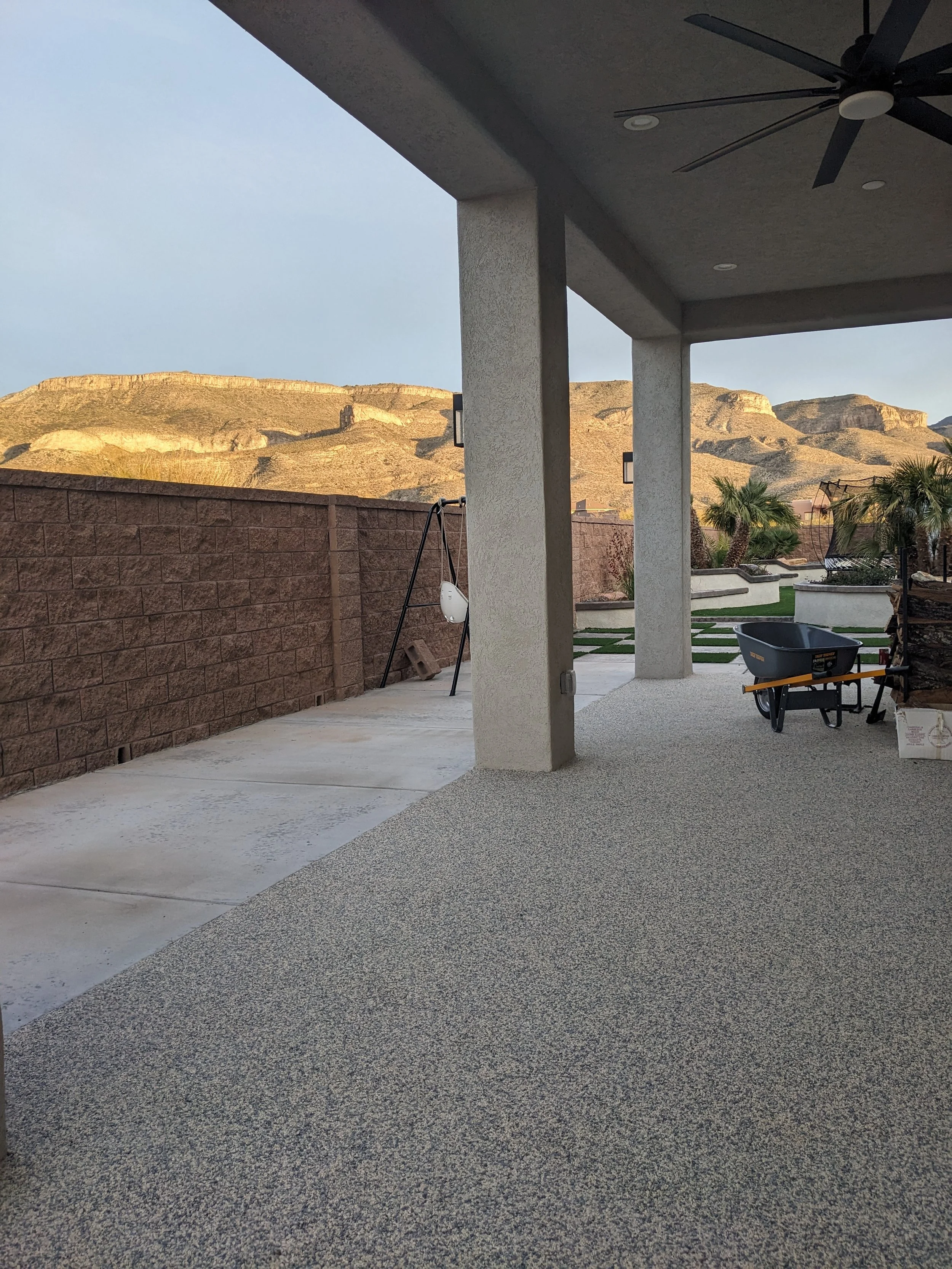 View of an outdoor patio area with a ceiling fan, a wheelbarrow, a brick wall, a mountain landscape in the background, and some palm trees.