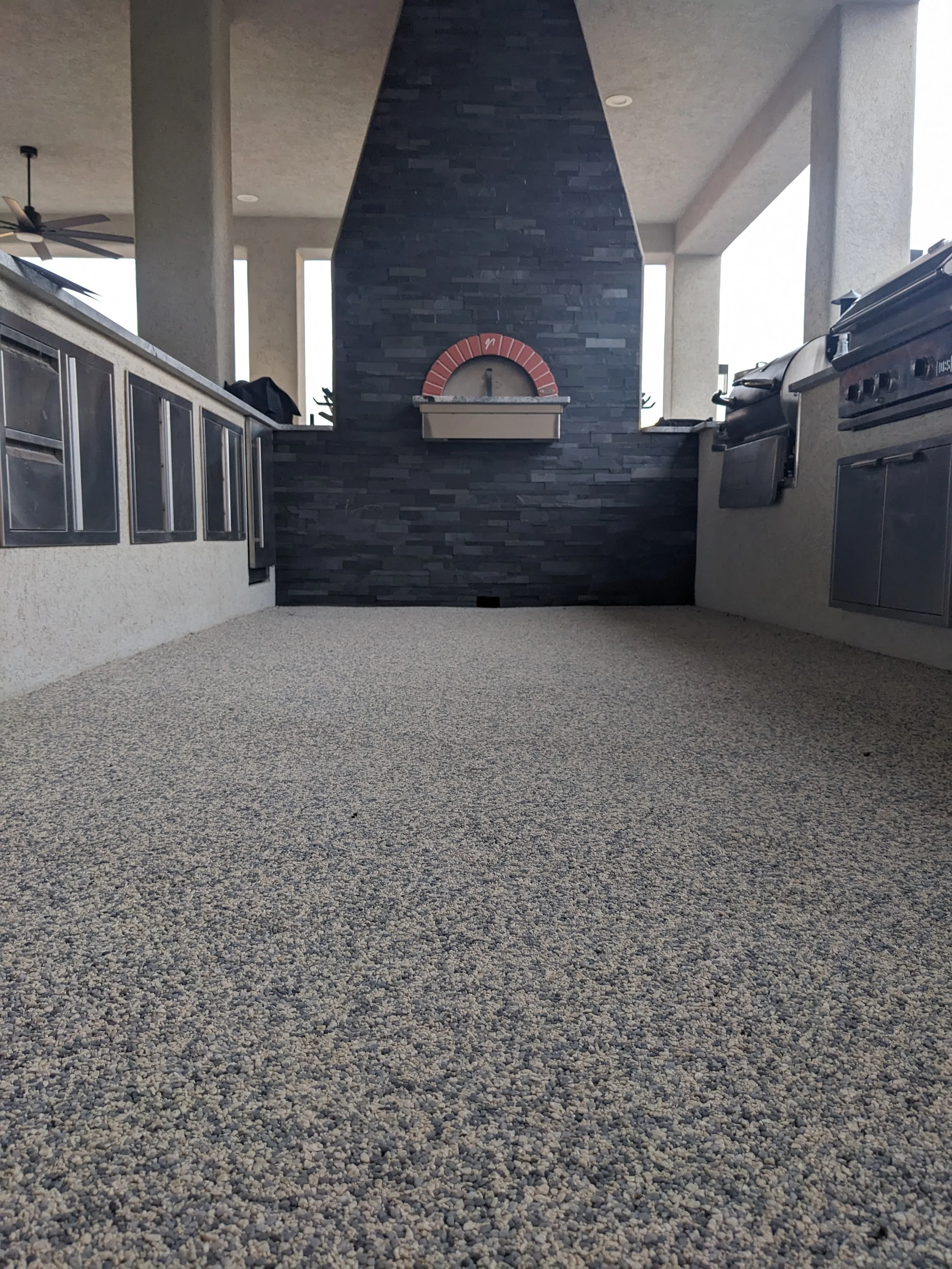 Outdoor kitchen with a black stone fireplace and stainless steel appliances, covered patio area with beige concrete columns and ceiling, ceiling fan, and window openings in the background.
