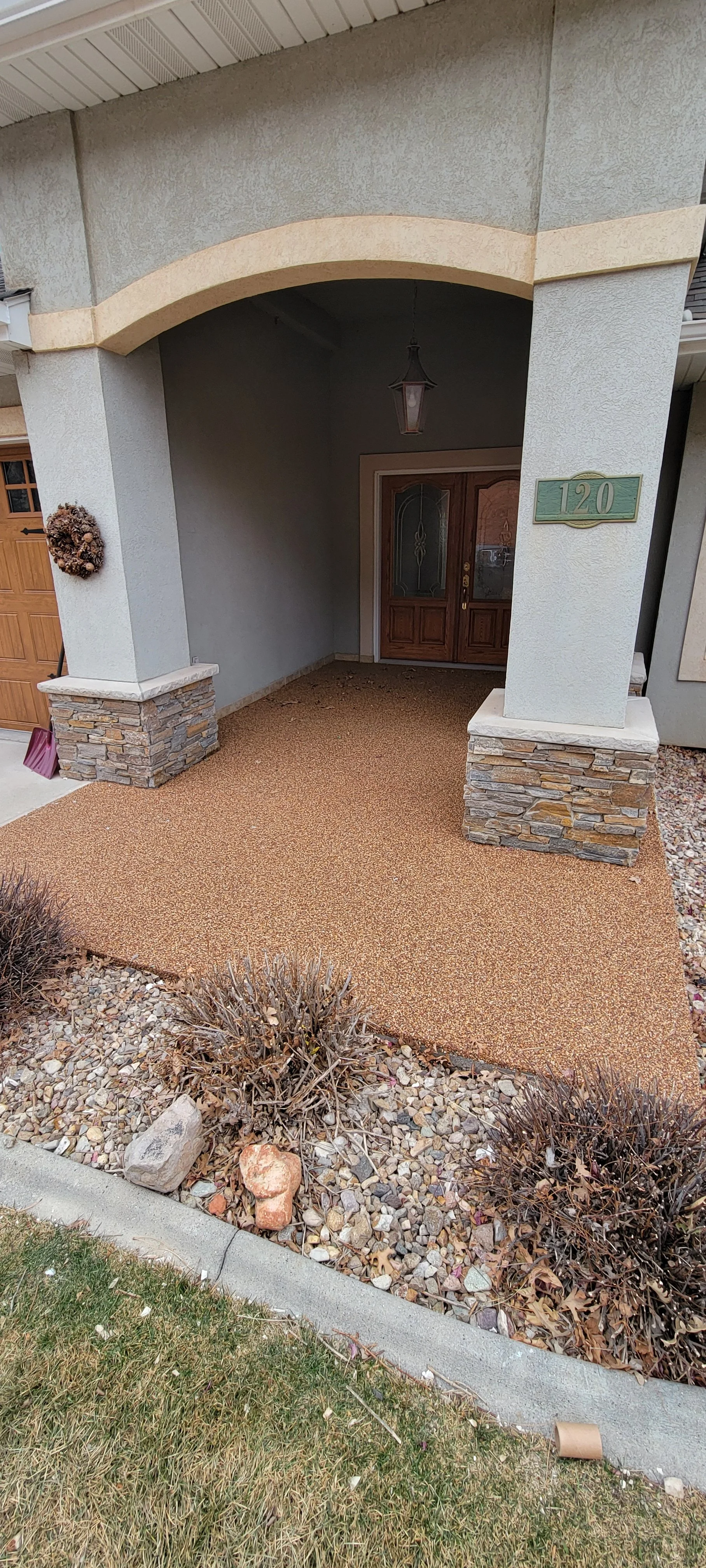 Front porch of a house with a stone foundation, stucco walls, a wooden front door, and a potted plant wreath, with a gravel and lawn area in front.