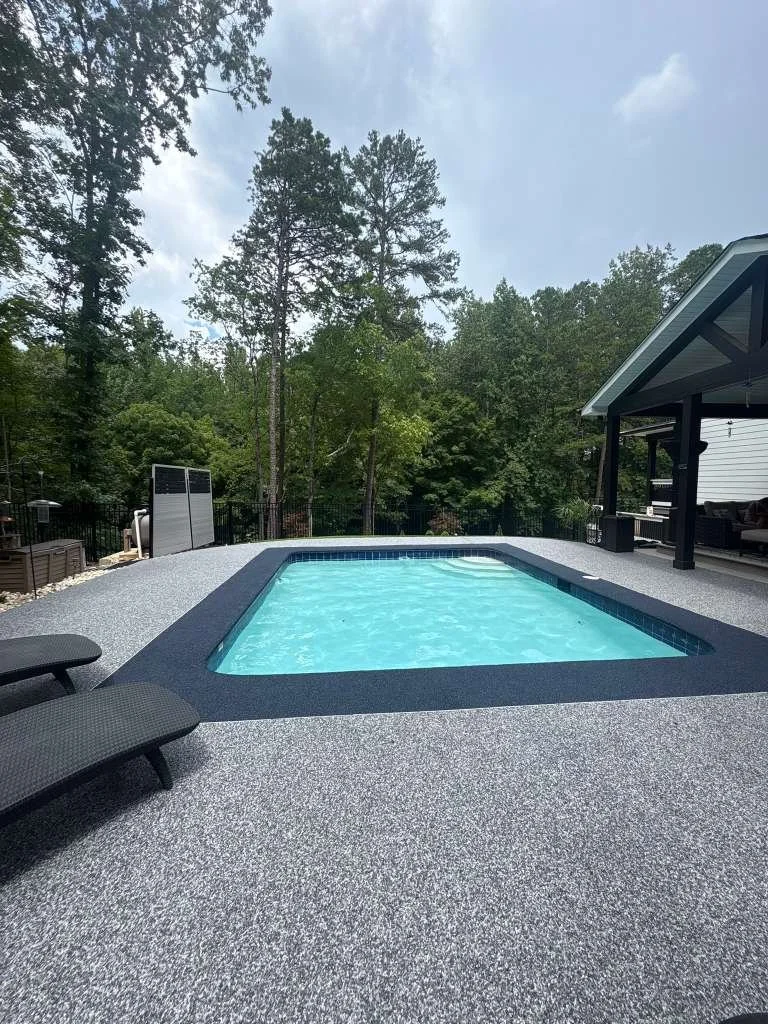 A backyard with a small blue swimming pool surrounded by a textured gray pool deck, with two black lounge chairs on the left, and tall trees in the background under cloudy skies.