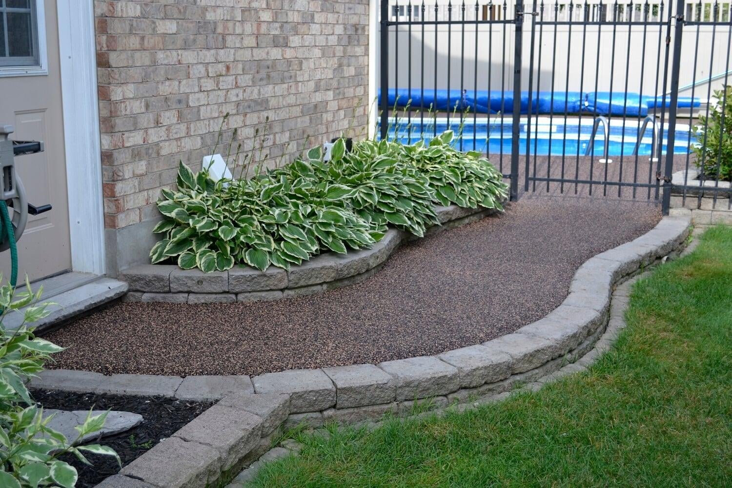 Garden bed with green and white variegated hosta plants, bordered by gray stone blocks, adjacent to a house with brick exterior, and a black metal gate leading to a swimming pool.