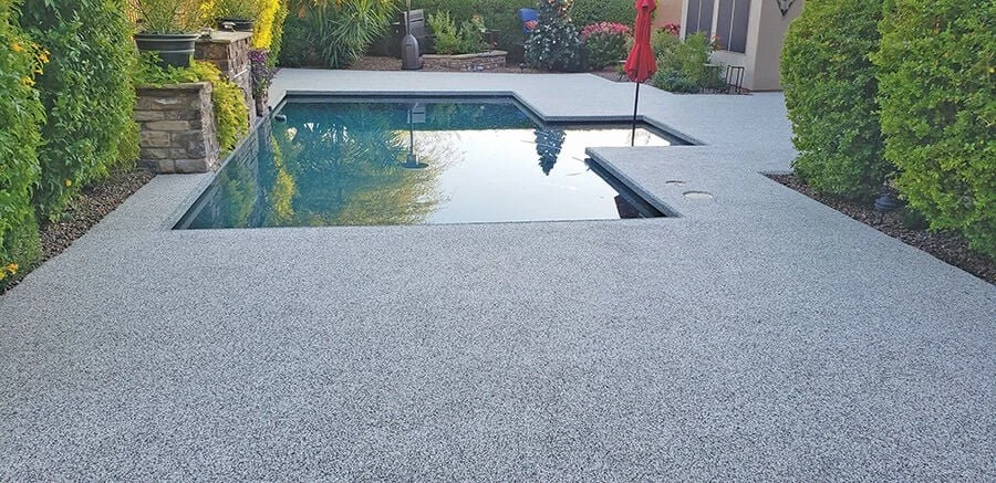 Backyard with a rectangular swimming pool, surrounded by a textured concrete deck, with trees and bushes on the sides and a patio area with a red umbrella in the background.
