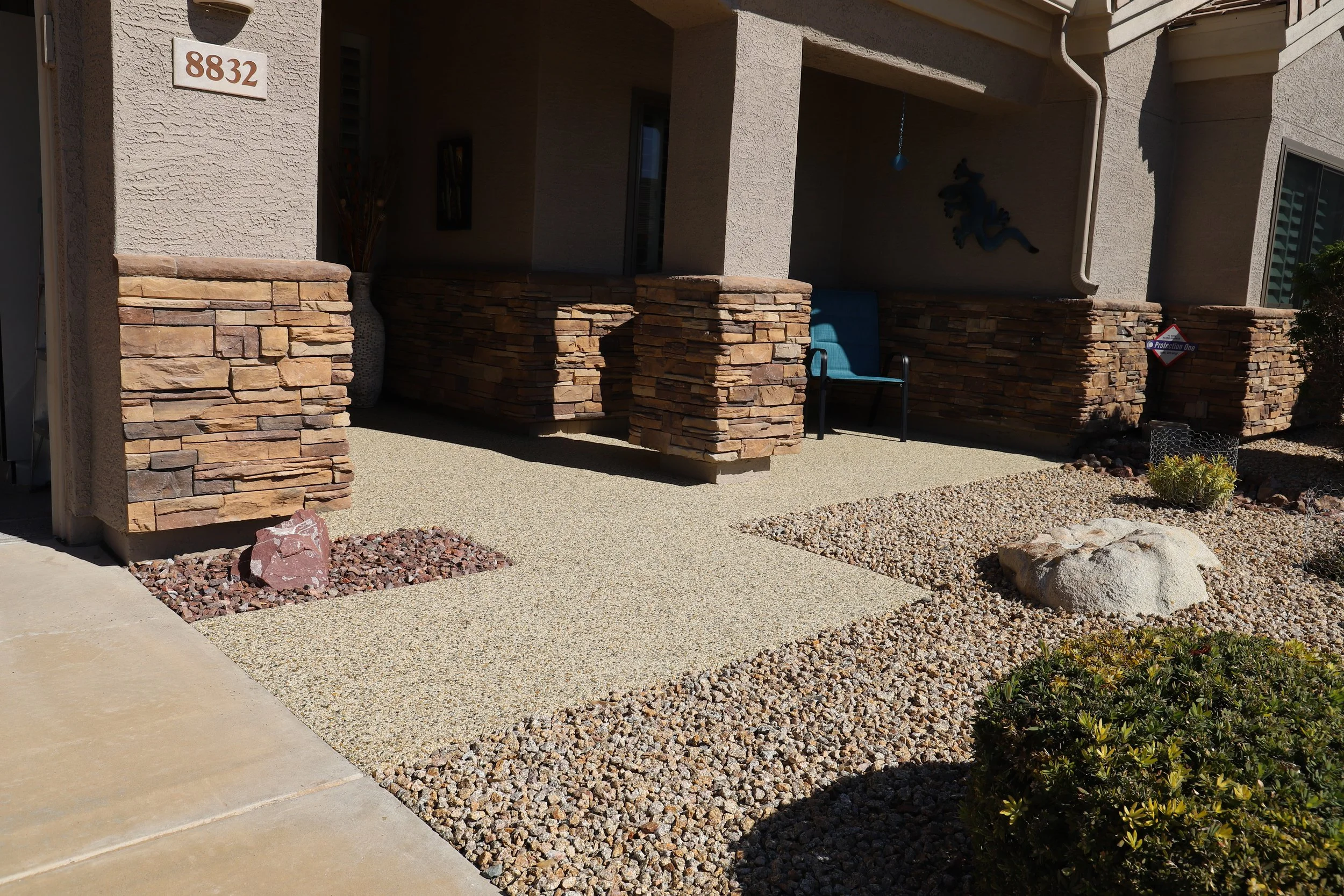 The front porch of a house with beige stucco walls, brick pillars, decorative stonework, and gravel landscaping. There is a blue chair and a wall-mounted gecko decoration on the porch.