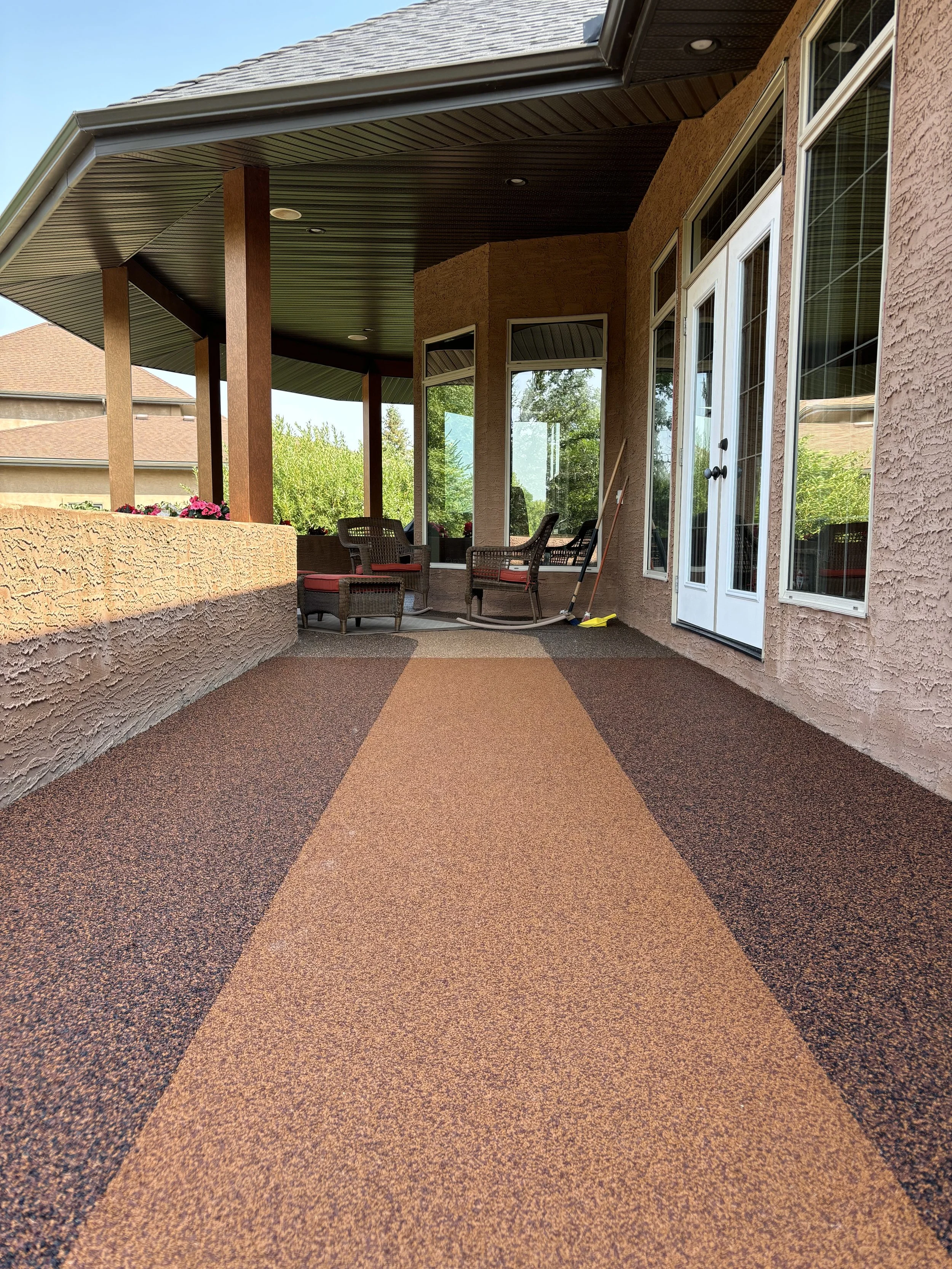 A covered patio area with a textured, two-tone flooring, outdoor furniture including chairs with red cushions, potted flowers, and a broom leaning against the wall. The patio is attached to a house with large windows and a glass door, with a view of 
