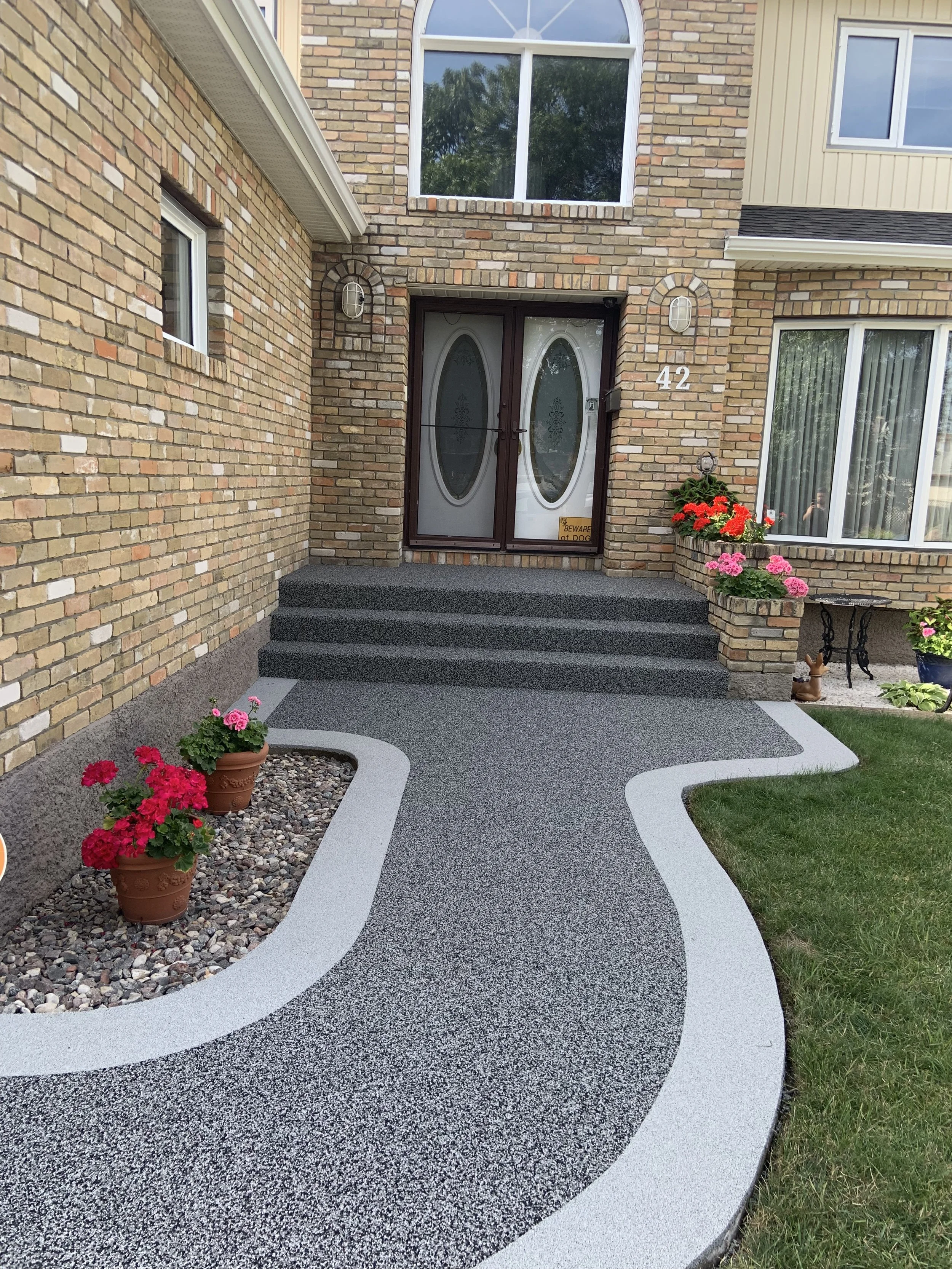 A brick house with a double front door, decorated with potted plants and flowers. The pathway leading to the door has a patterned design with curving lines.