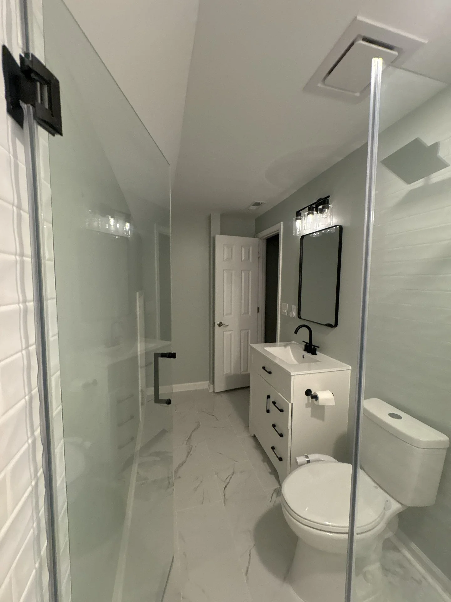 Modern bathroom with gray walls, marble floor, white vanity with black faucet, wall-mounted mirror, black lighting fixture, glass shower door, and toilet.