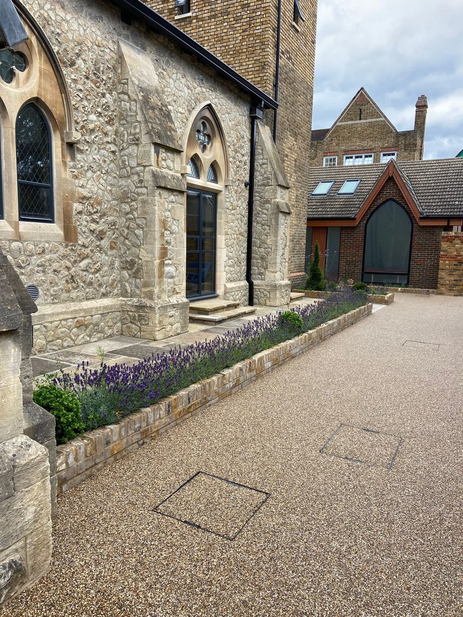 Exterior view of a stone and brick building with arched windows, a garden bed with purple flowers, and a textured gravel walkway.