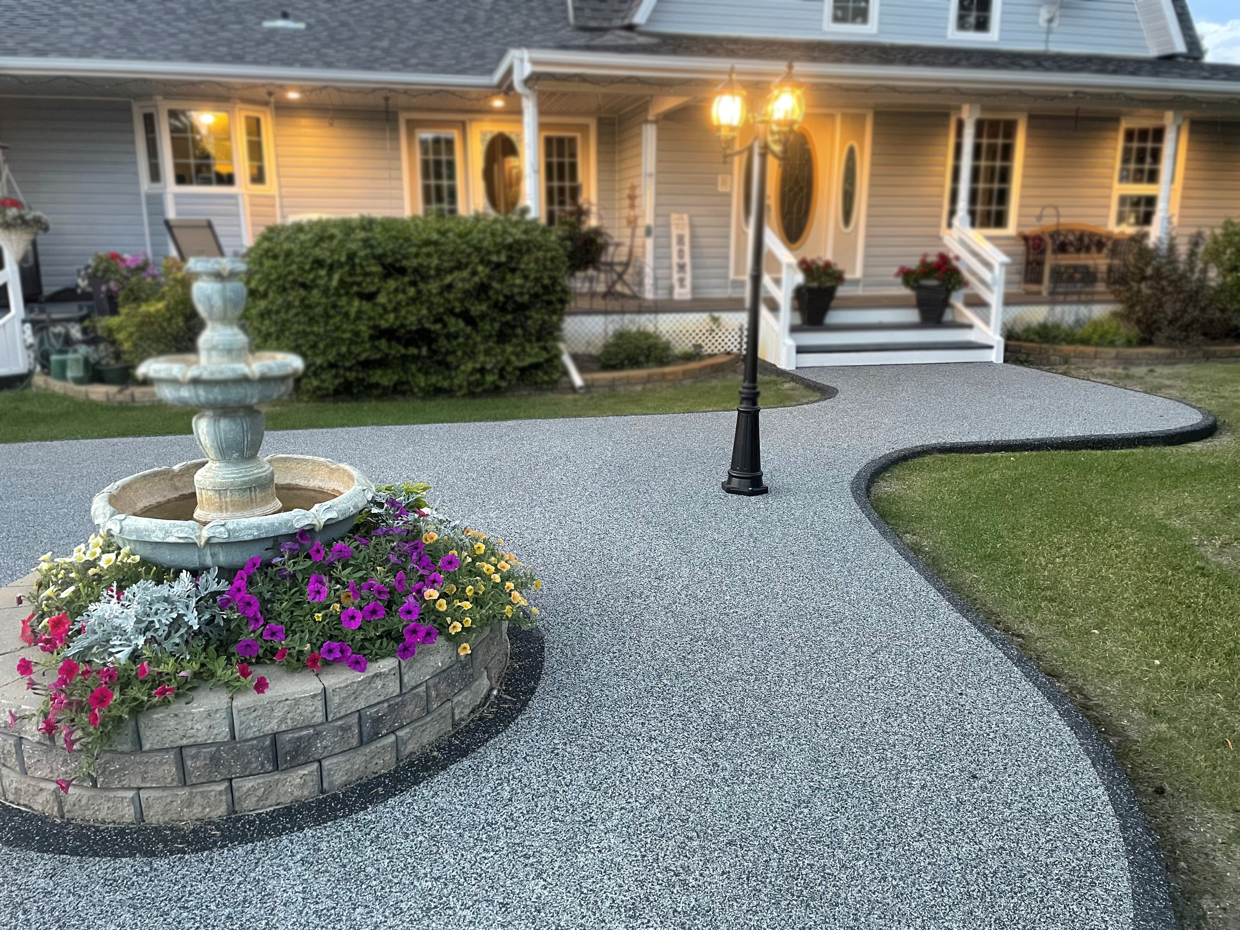 Front yard of a house with a concrete path, a flower bed with colorful flowers, a fountain, a lamppost, and a porch with stairs and potted plants.