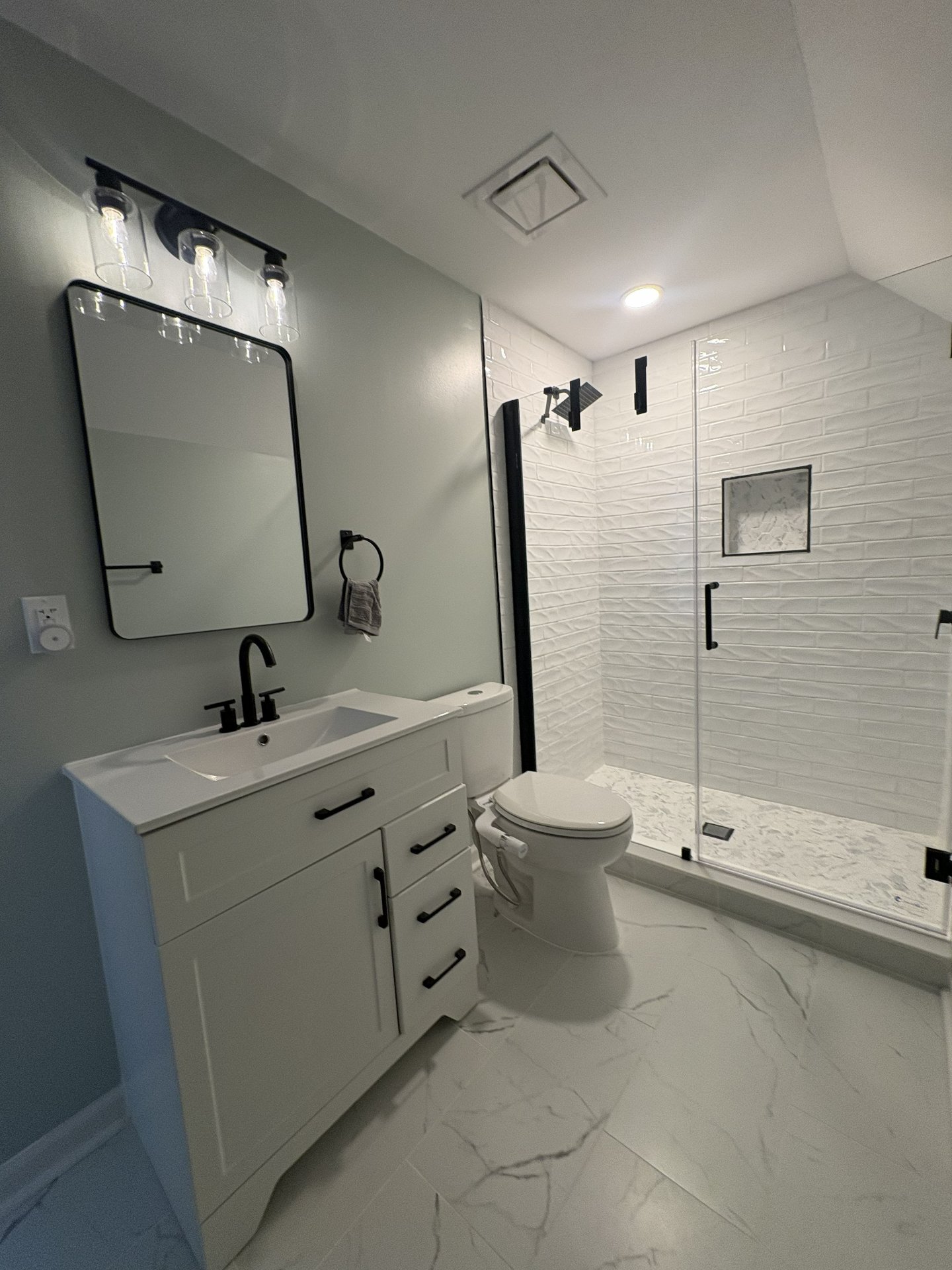 Modern bathroom with white vanity, black fixtures, mirror, toilet, and a walk-in shower with glass doors, white textured tiles, and black fixtures.