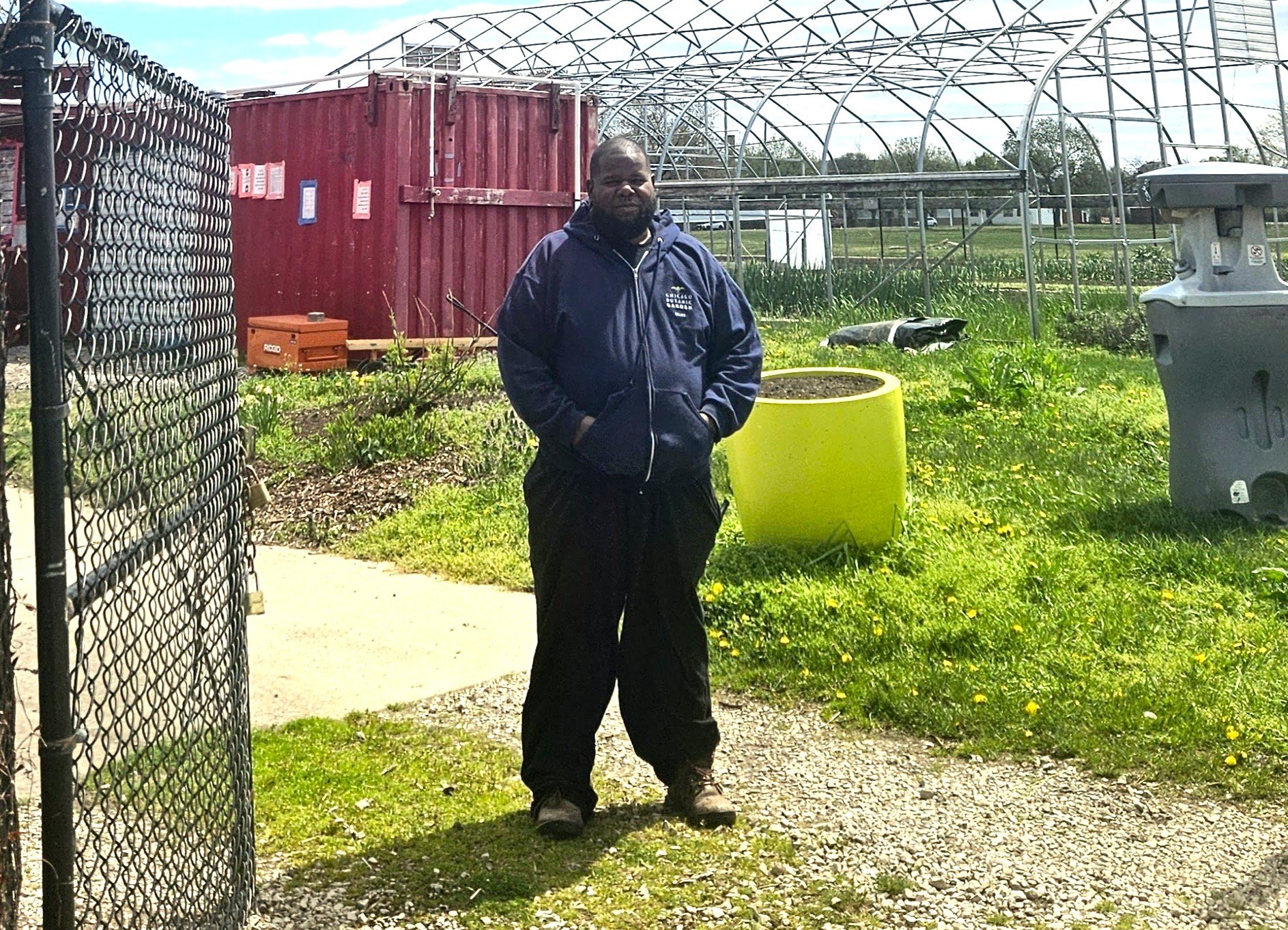 A man standing outdoors near a chain-link fence, with a red container, a yellow planter, and a greenhouse structure in the background on a sunny day.