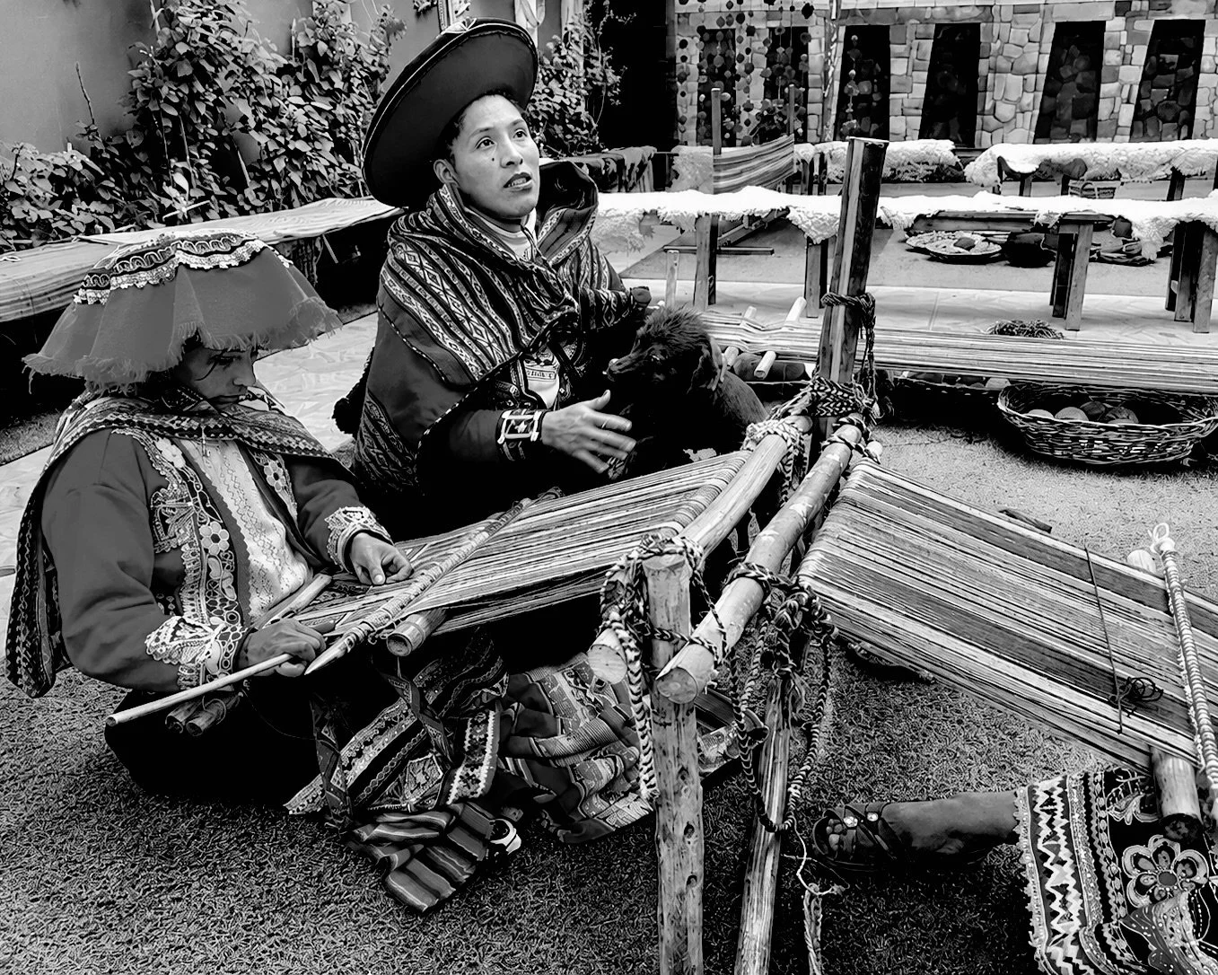 Two women in traditional clothing, one with a large hat, are weaving on a loom while sitting on the ground, with various woven textiles and supplies around them.