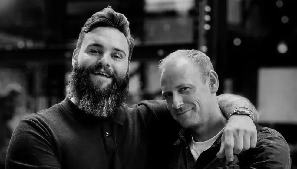 Two men smiling, one with a beard and the other clean-shaven, standing close together with the man on the right having his arm around the other, in an indoor setting.