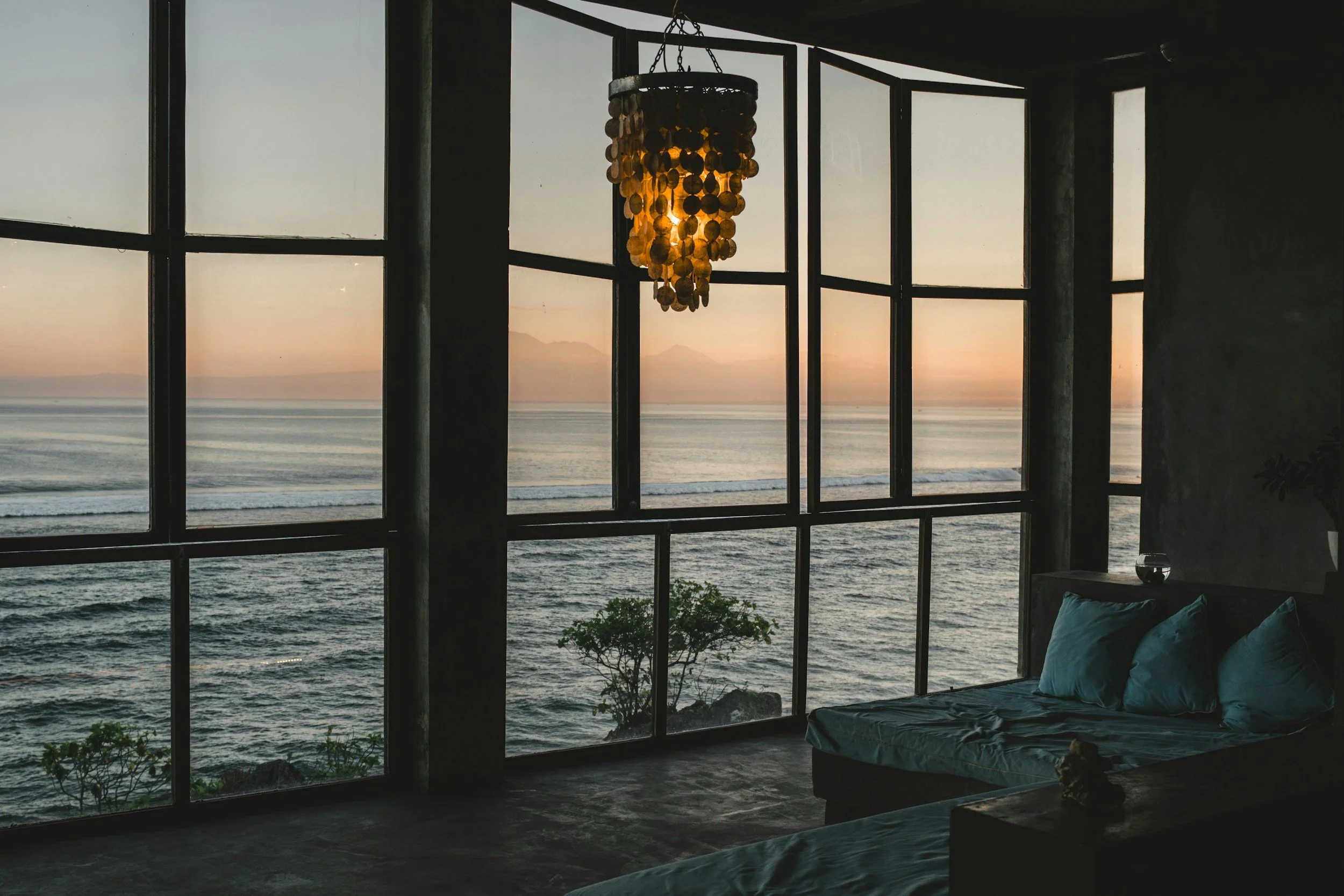 View of the ocean at sunset through large glass windows in a room. A chandelier hangs from the ceiling, and part of a bed with blue pillows is visible.