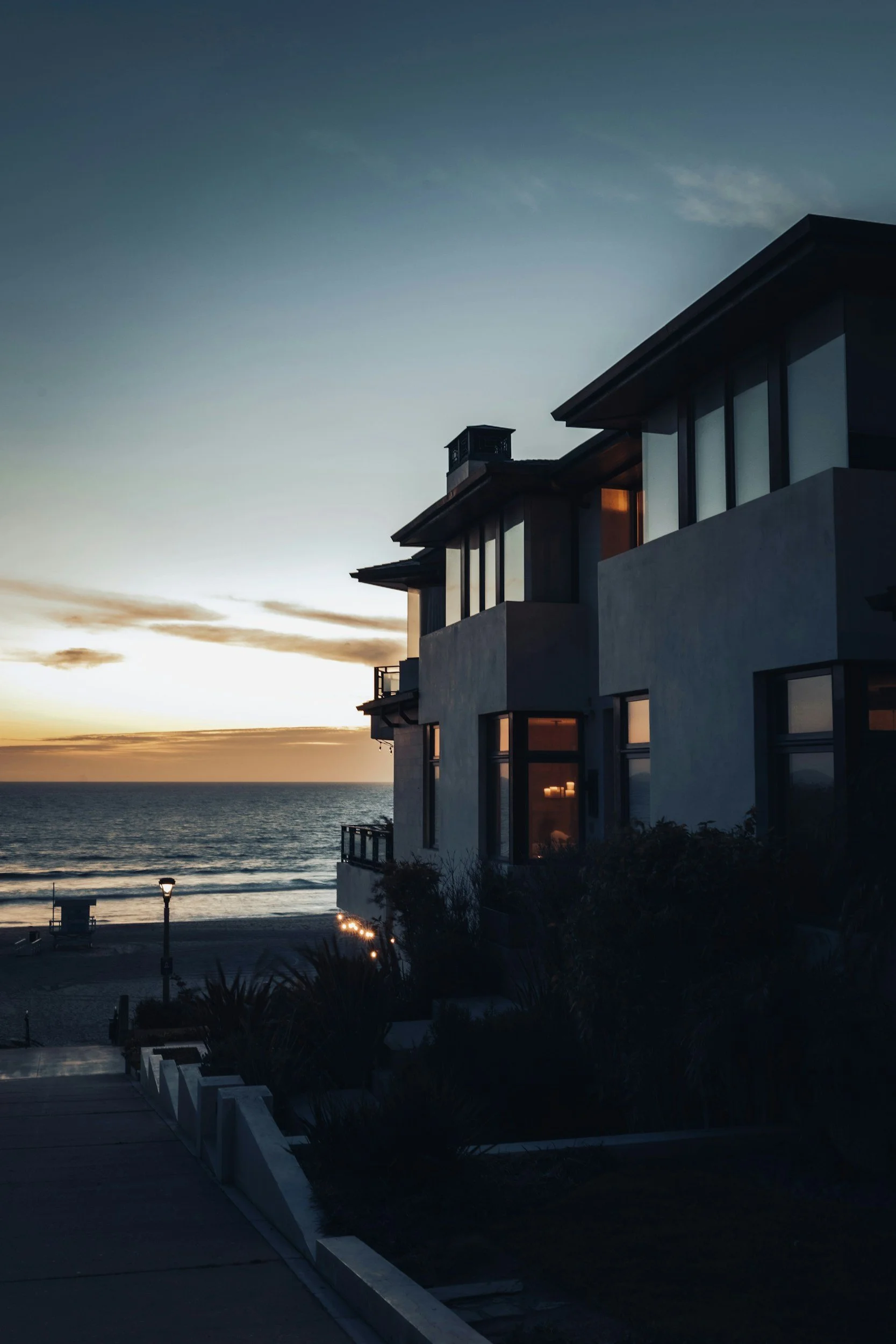Modern multi-story house with large windows overlooking the ocean at sunset, with a pathway and some plants in the foreground.