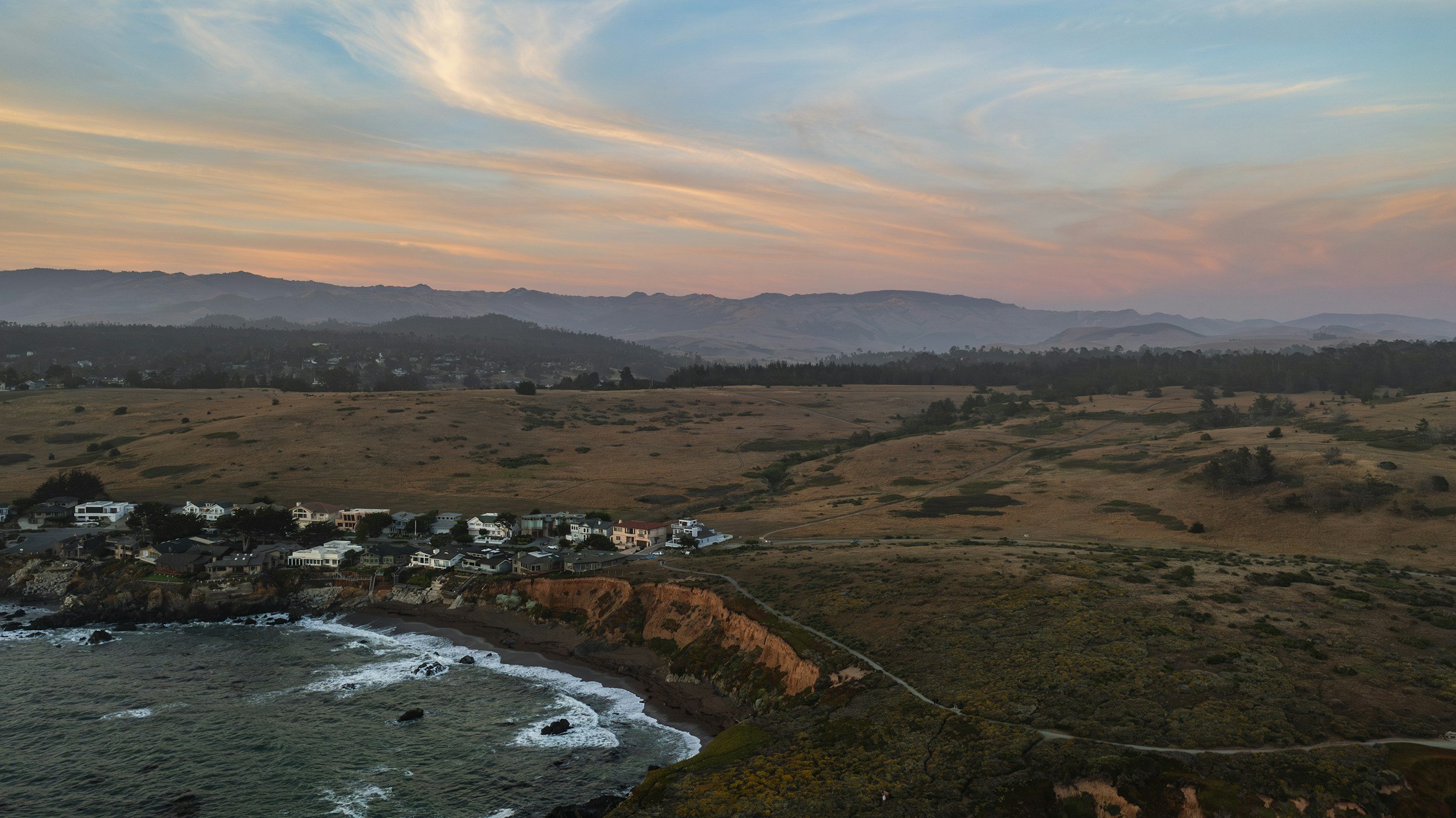 Scenic coastal landscape at sunset with rolling hills, houses near the shoreline, and a partly cloudy sky with pastel hues.