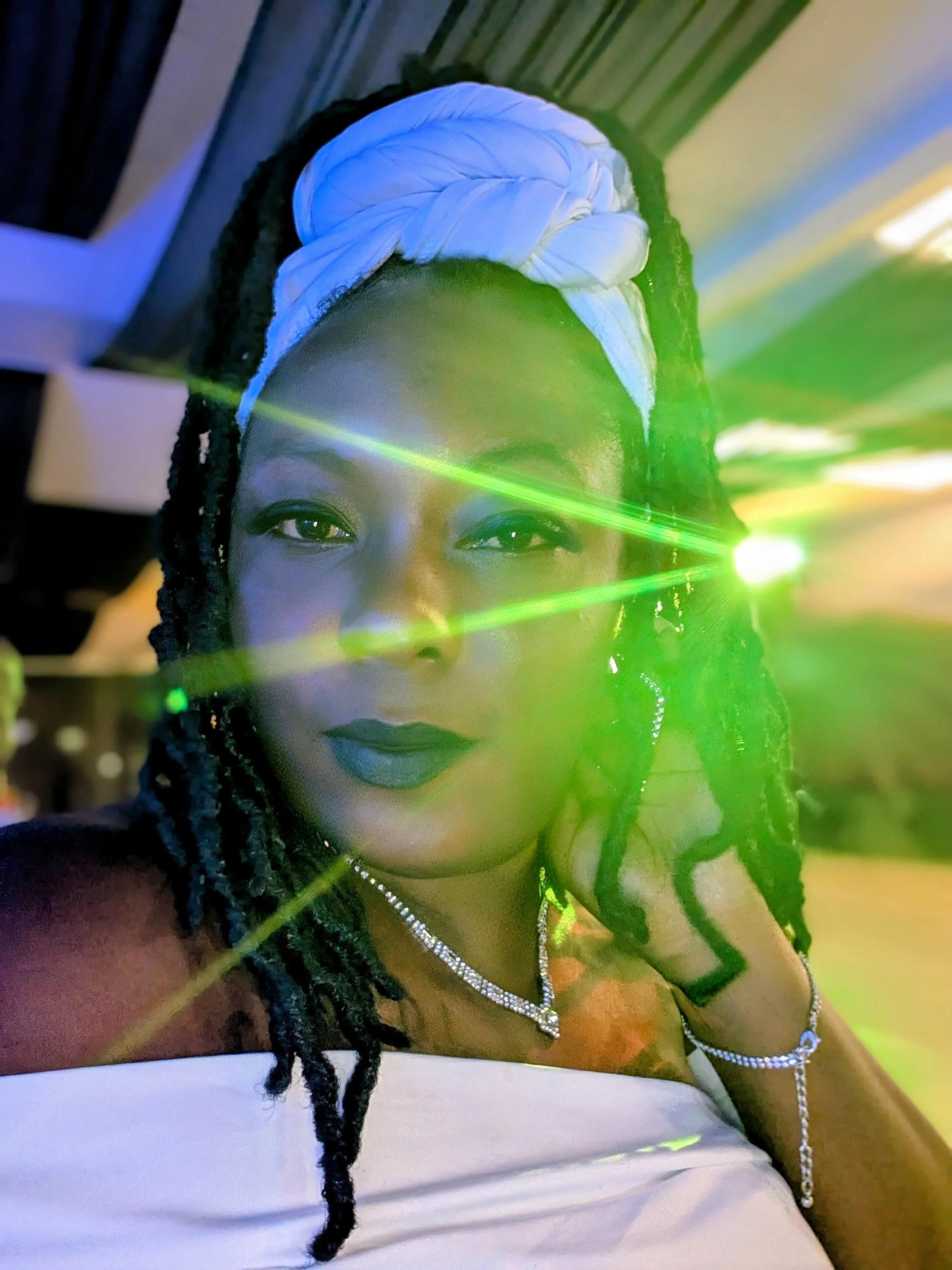A woman with dark braided hair wearing a white headwrap, necklace, and earrings, posing indoors with green laser light reflections.