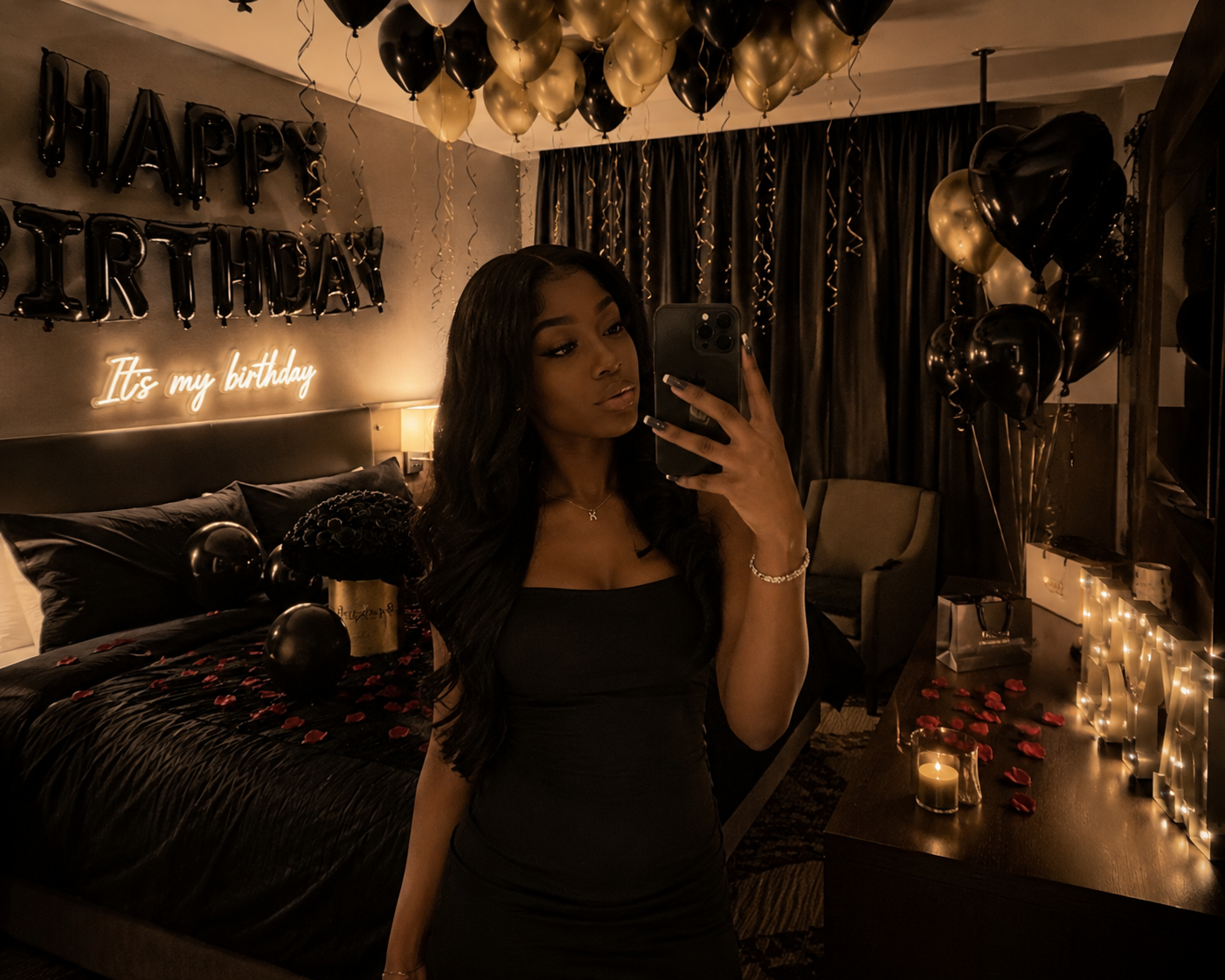 A woman taking a selfie in a bedroom decorated for a birthday celebration, with black and gold balloons, a balloon spelling out "HAPPY BIRTHDAY," and a neon sign that reads "It's my birthday." Rose petals and candles are on the bed and dresser.