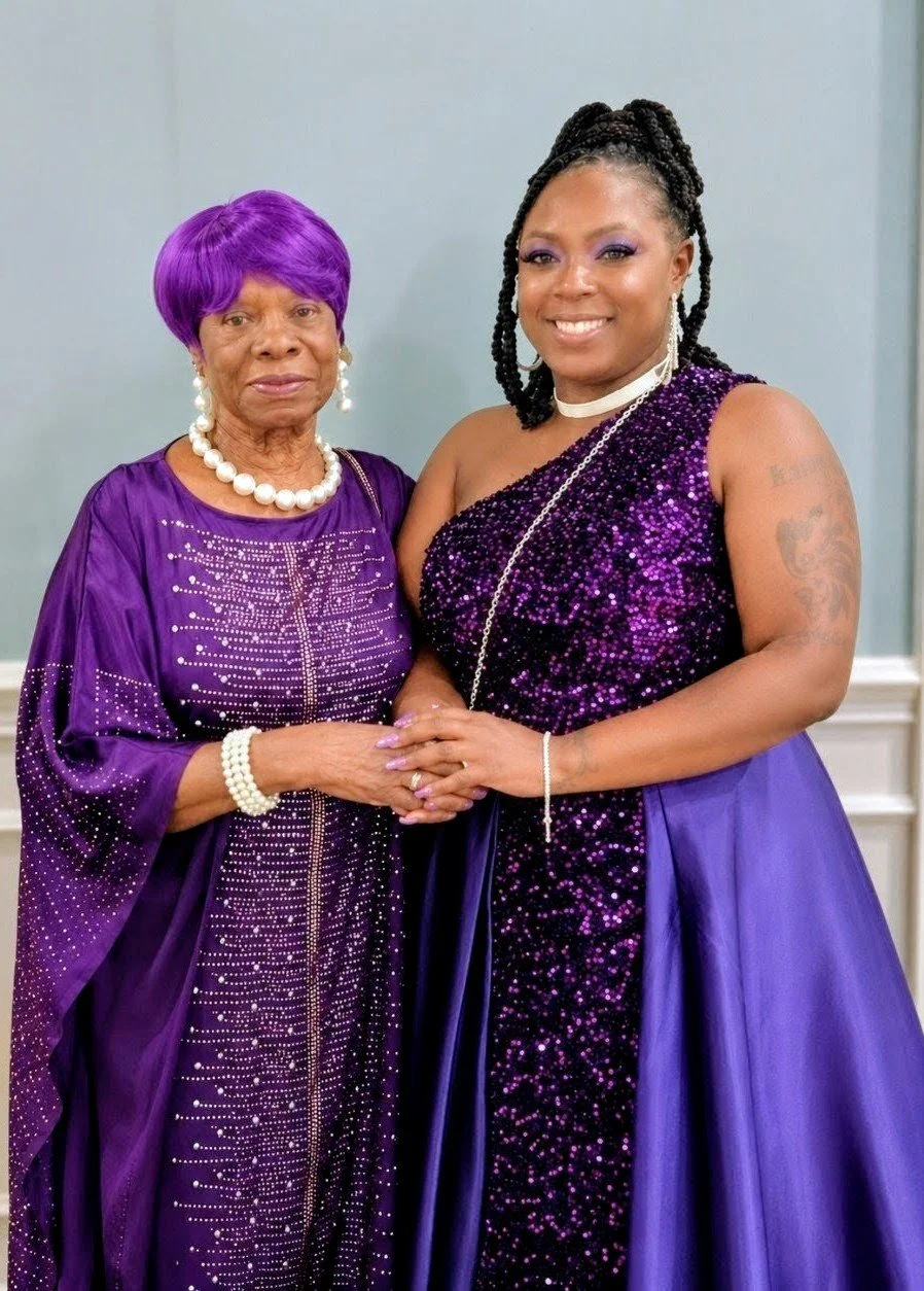 Two women wearing purple dresses standing and holding hands, smiling, with a plain blue wall background.