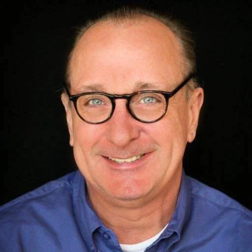 Close-up portrait of a smiling middle-aged man with glasses, wearing a blue shirt against a black background.