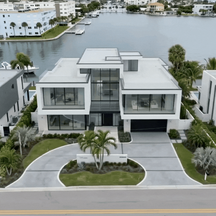 Drone orbit of modern waterfront house with large glass windows, front driveway, and lush landscaping.