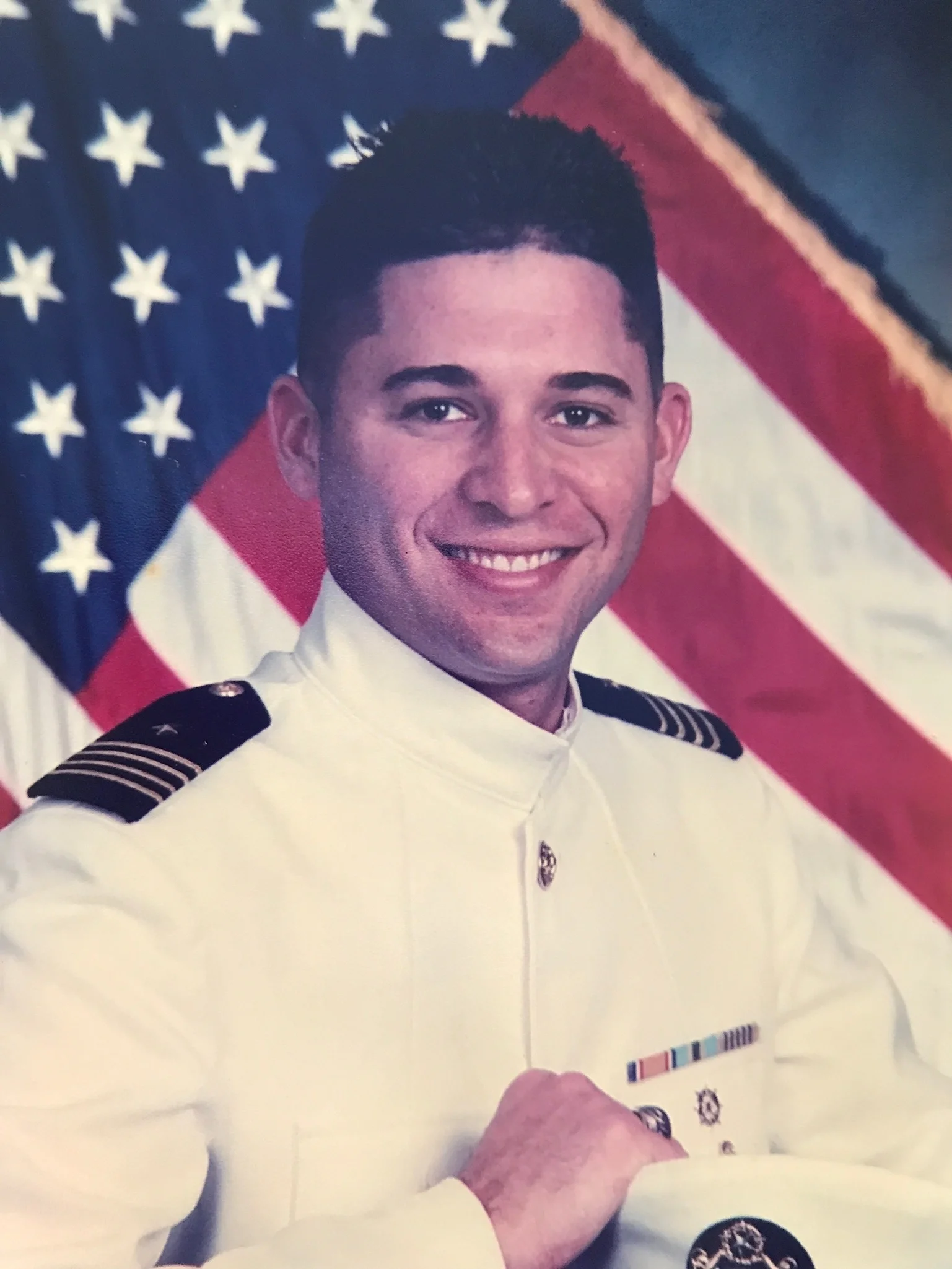 Young man in a dress whites, a white military uniform with naval insignia, smiling, with an American flag in the background.