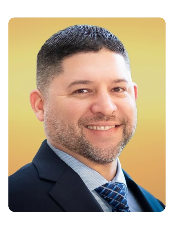 A smiling man in a business suit with a blue tie, short dark hair, and facial hair, against a yellow background.