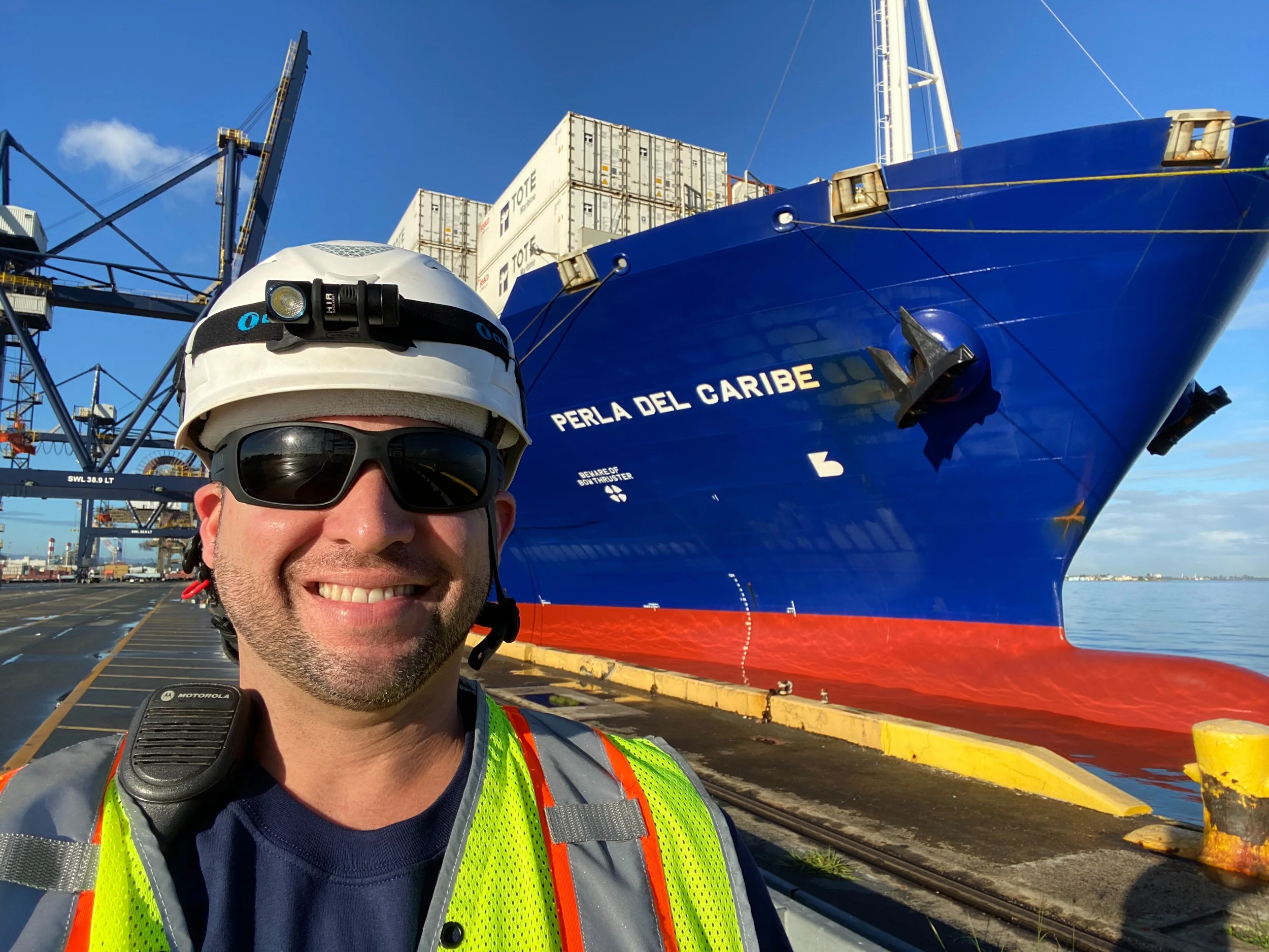 A worker wearing a white safety helmet, sunglasses, and a high-visibility yellow and gray safety vest takes a selfie in front of a large cargo ship named 'Perla del Caribe' at a dock. The ship is blue with containers on deck, and industrial cranes are visible in the background under a clear blue sky.