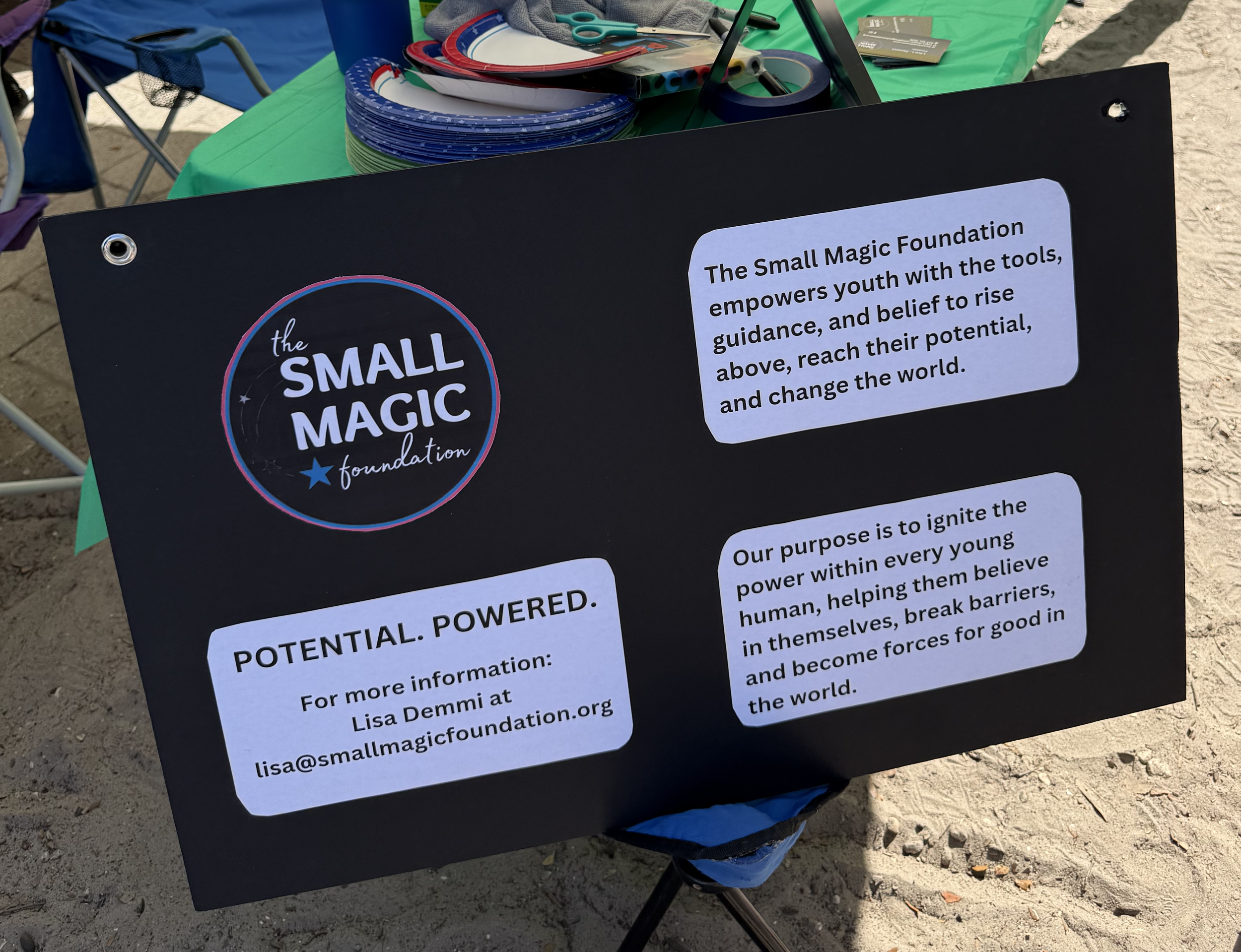 Black sign promoting the Small Magic Foundation with white text, placed on a table with colorful ribbons and other materials, outdoors on sandy ground.