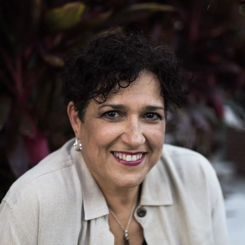 A woman with short, curly dark hair smiling outdoors, wearing a light-colored jacket and earrings, with blurred foliage in the background.
