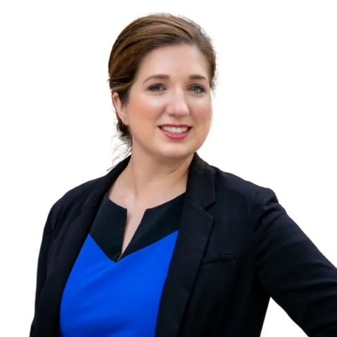 A woman with light brown hair in a ponytail, wearing a dark blazer over a blue top, smiling at the camera against a plain white background.