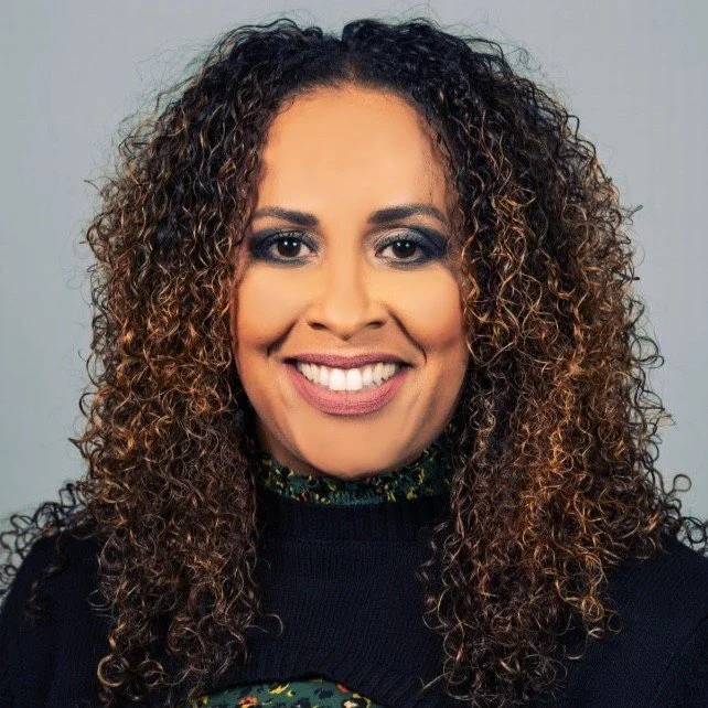 A woman with curly brown hair smiling at the camera against a gray background.