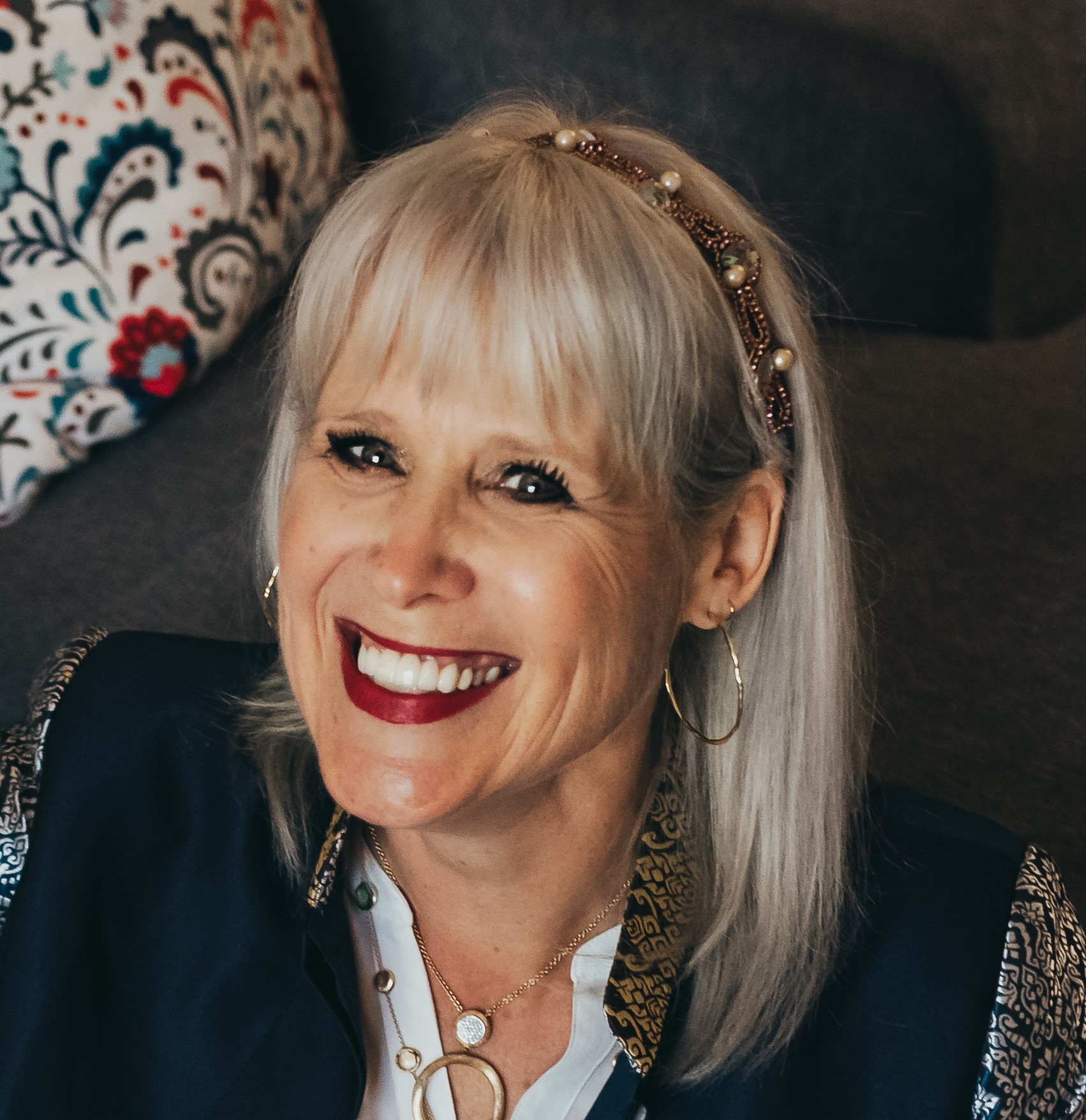 A happy woman with blonde hair, wearing a pearl and beaded headband, gold hoop earrings, a gold necklace, and a dark jacket with patterned sleeves, smiling in a cozy setting with pillows in the background.