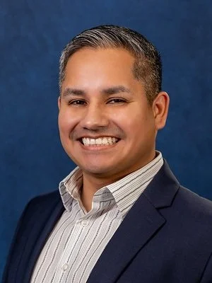 A smiling man in a suit and striped shirt against a blue background.