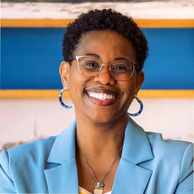 Smiling woman with short curly hair, glasses, hoop earrings, and a blue blazer.